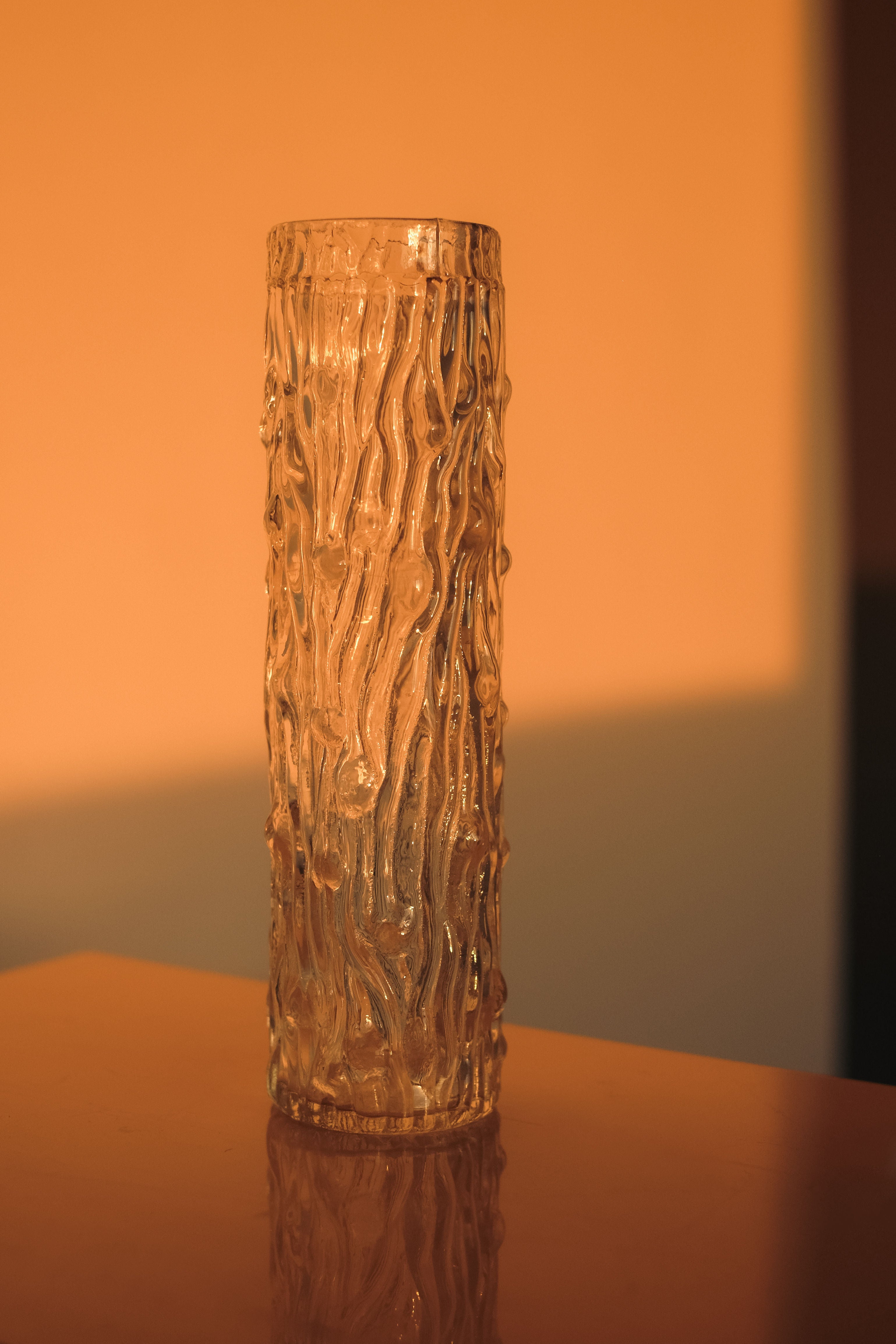 A small, textured septembre studios glass cylinder vase rests on a smooth surface under warm golden light. The bark-like patterns reflect the light, casting soft shadows. The background transitions from orange to cream, evoking a sunset.