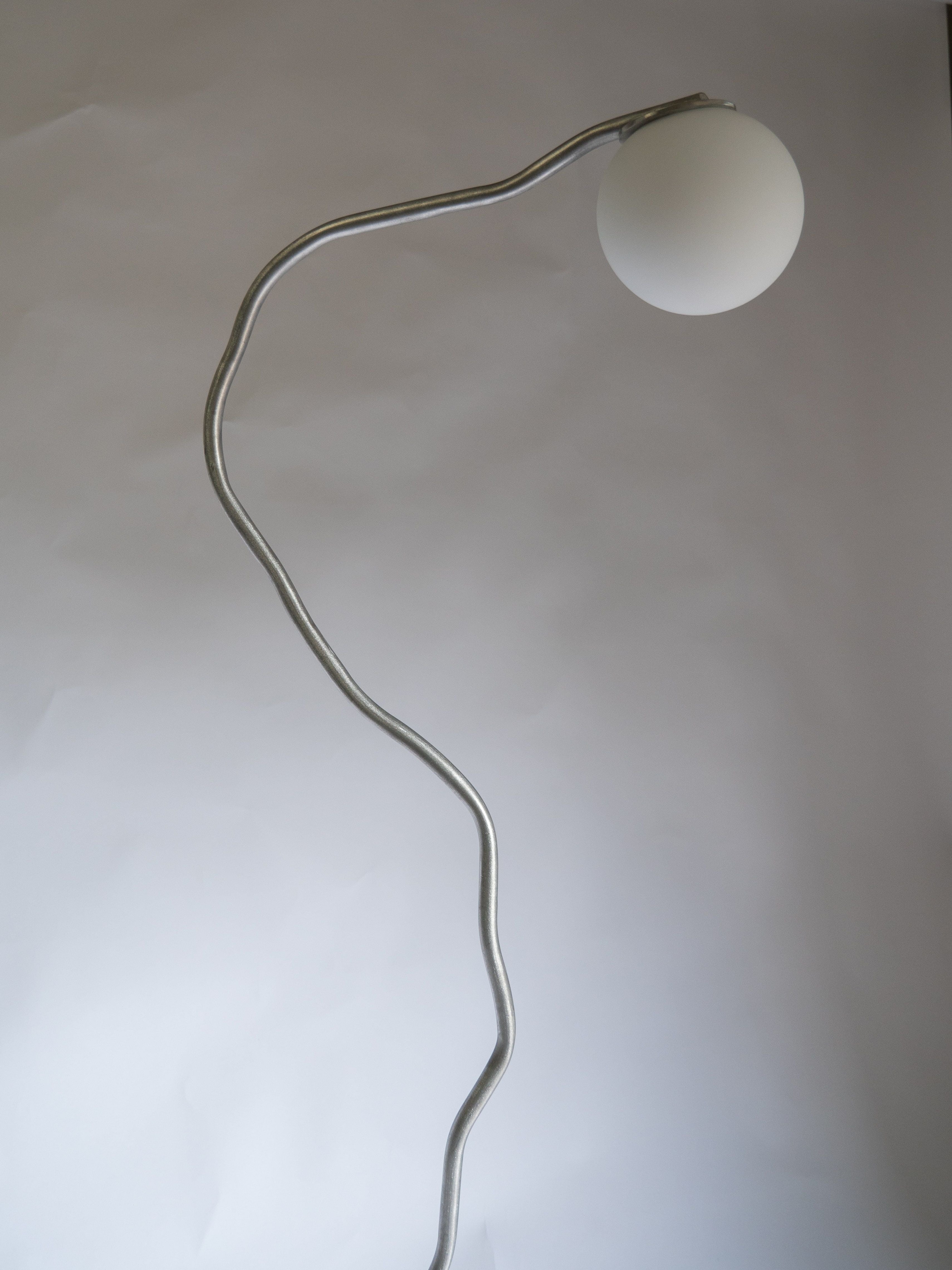 A modern Pea Head Floor Lamp by Six Dots Design with a thin, wavy metal stand and a round, white lampshade at the top, set against a plain, light-colored background. This free-form lamp has a minimalist design with an artistic, curving structure that adds natural ambience to any space.