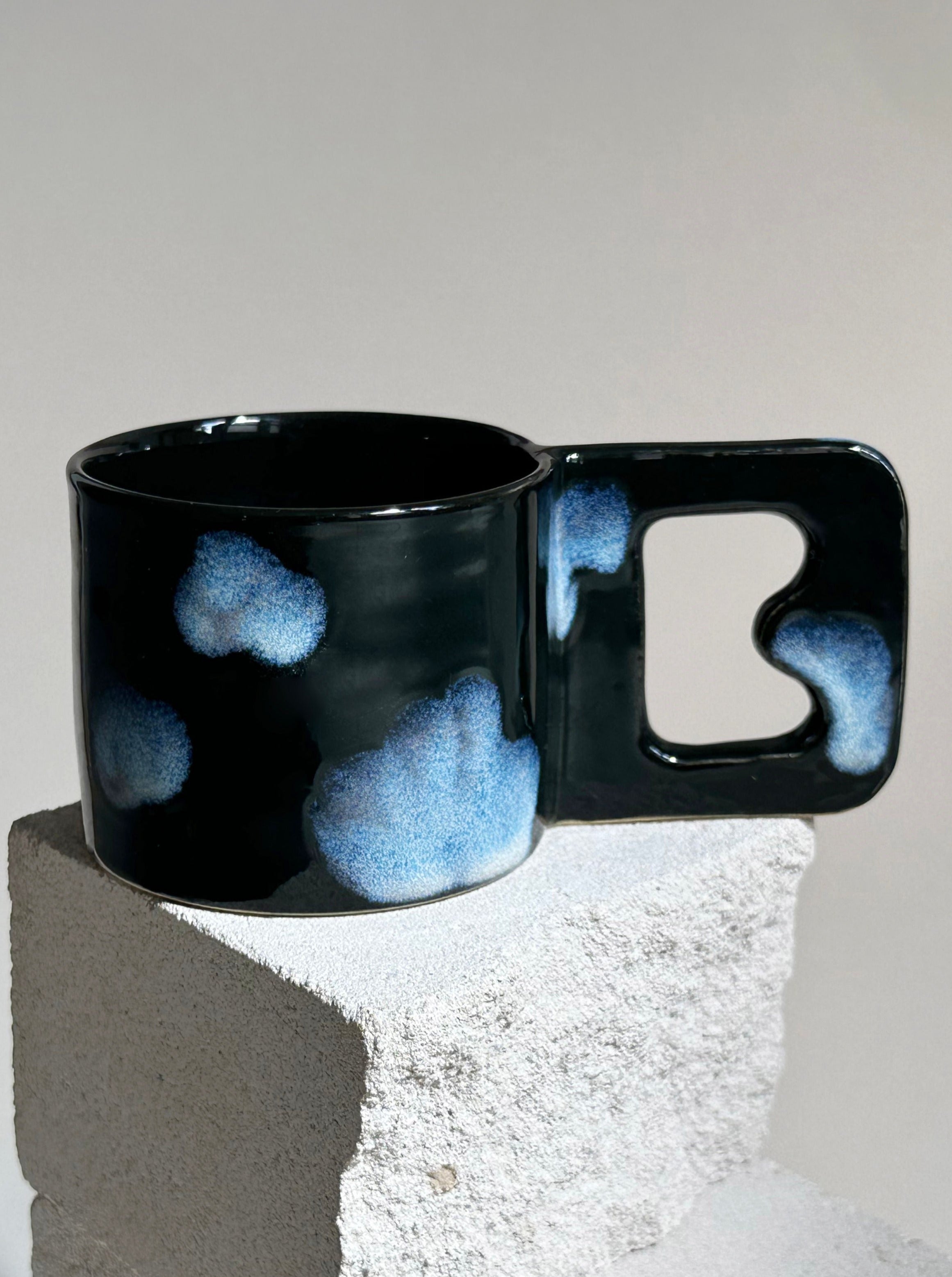 A dark-colored ceramic Cloud Cup by noki ceramics with blue cloud-like patterns on its surface is perched on a textured concrete block. Crafted from stoneware clay and adorned with food-safe glazes, the mug features a unique, angular handle with an open middle section. The minimalistic background highlights its artistic design.