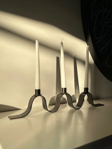Three white taper candles in Maud Vaughan’s handmade Ribbon Candelabra sit on a white surface, sunlight casting gentle shadows for a warm, minimalist effect.