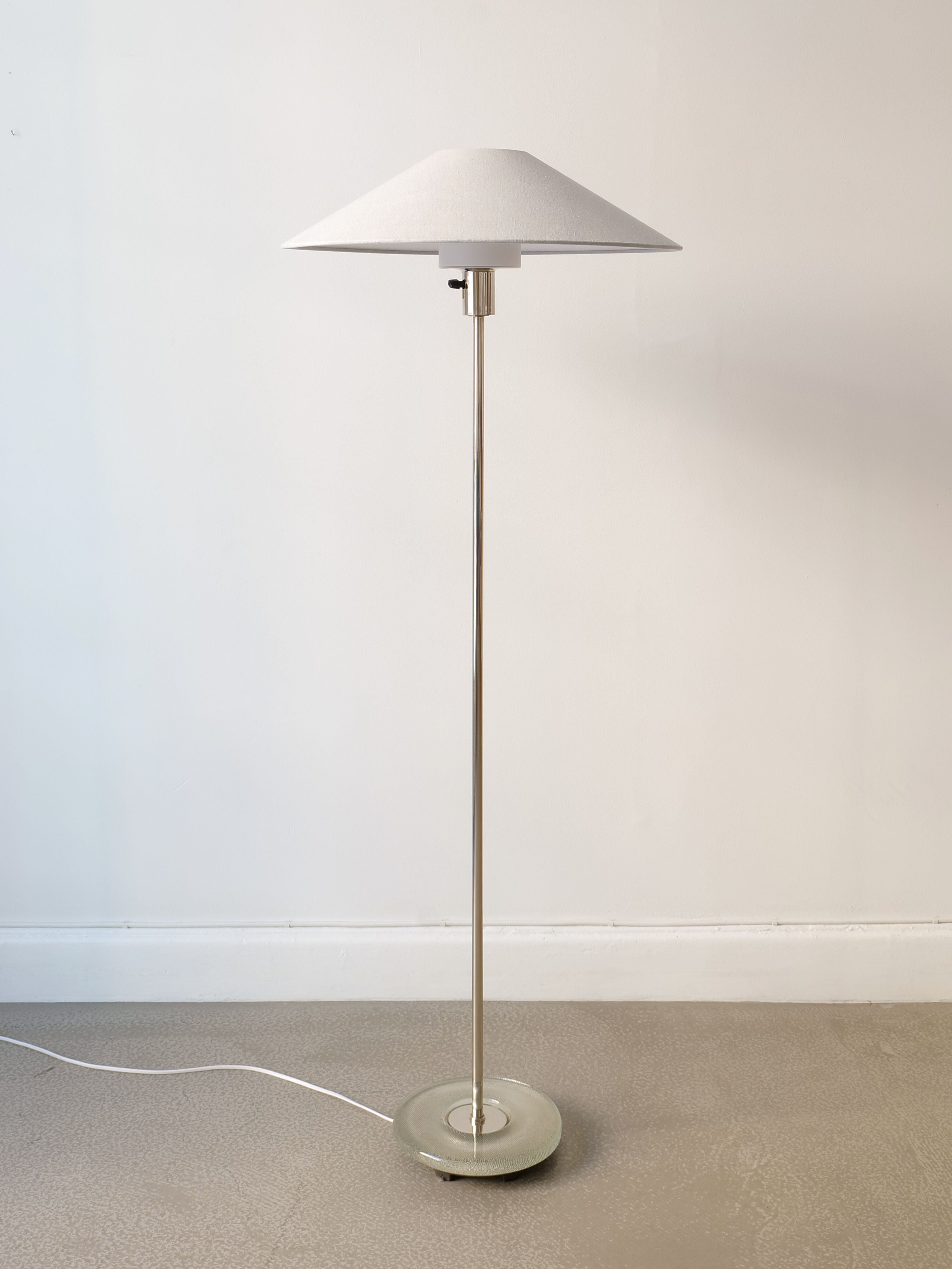 A sleek, modern floor lamp with a slim metallic stand and a cone-shaped white lampshade embodies the Swedish Modern design. This Deco Floor Lamp Harald Notini 1940s by Collection apart stands on a beige floor against a plain white wall, with a visible white power cord trailing from the base.
