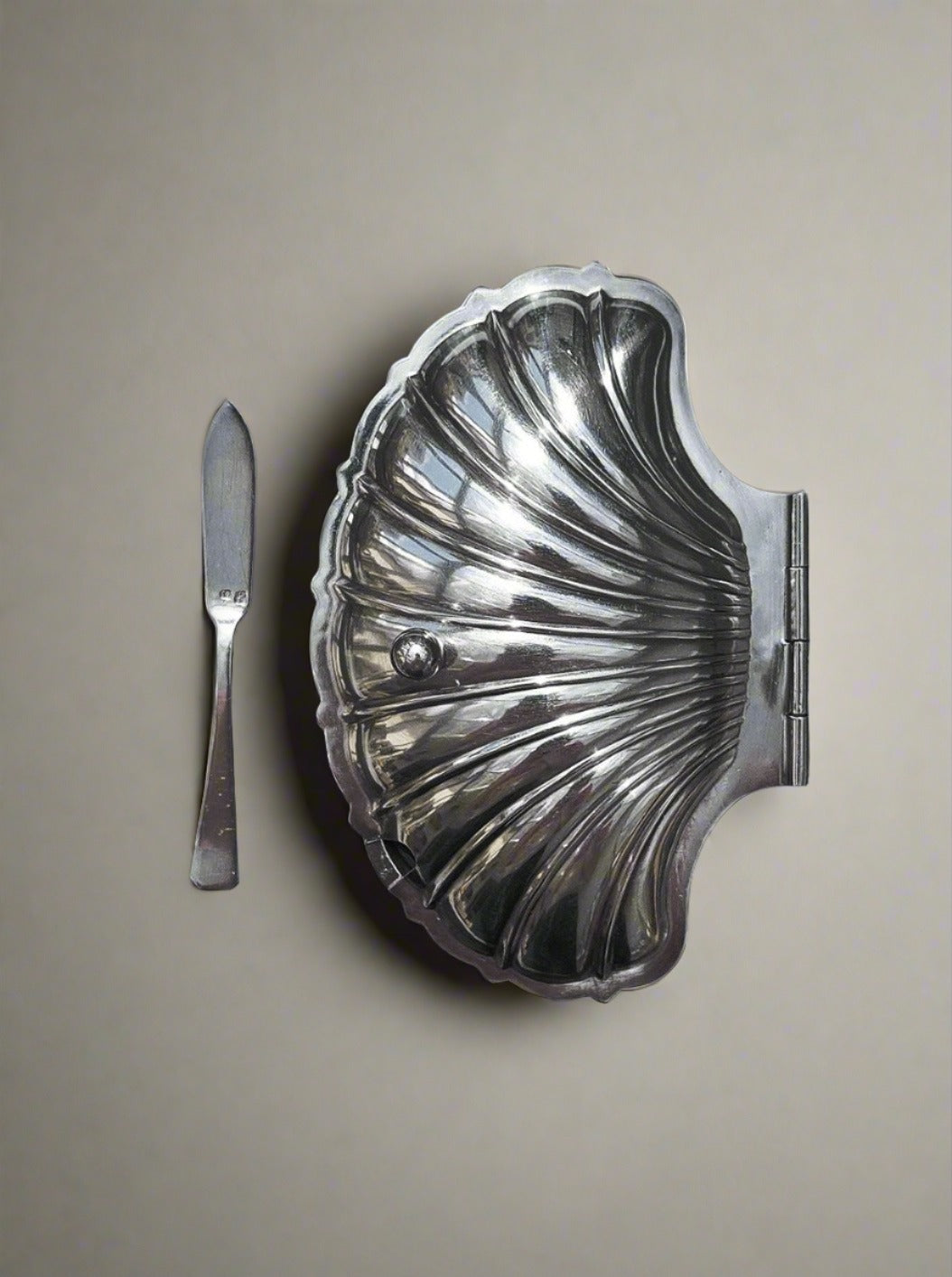 The Les Objoies Shell Silver-Plated Butter Dish exudes vintage charm with its hinged lid, placed open to the right of a matching silver butter knife on a plain background.