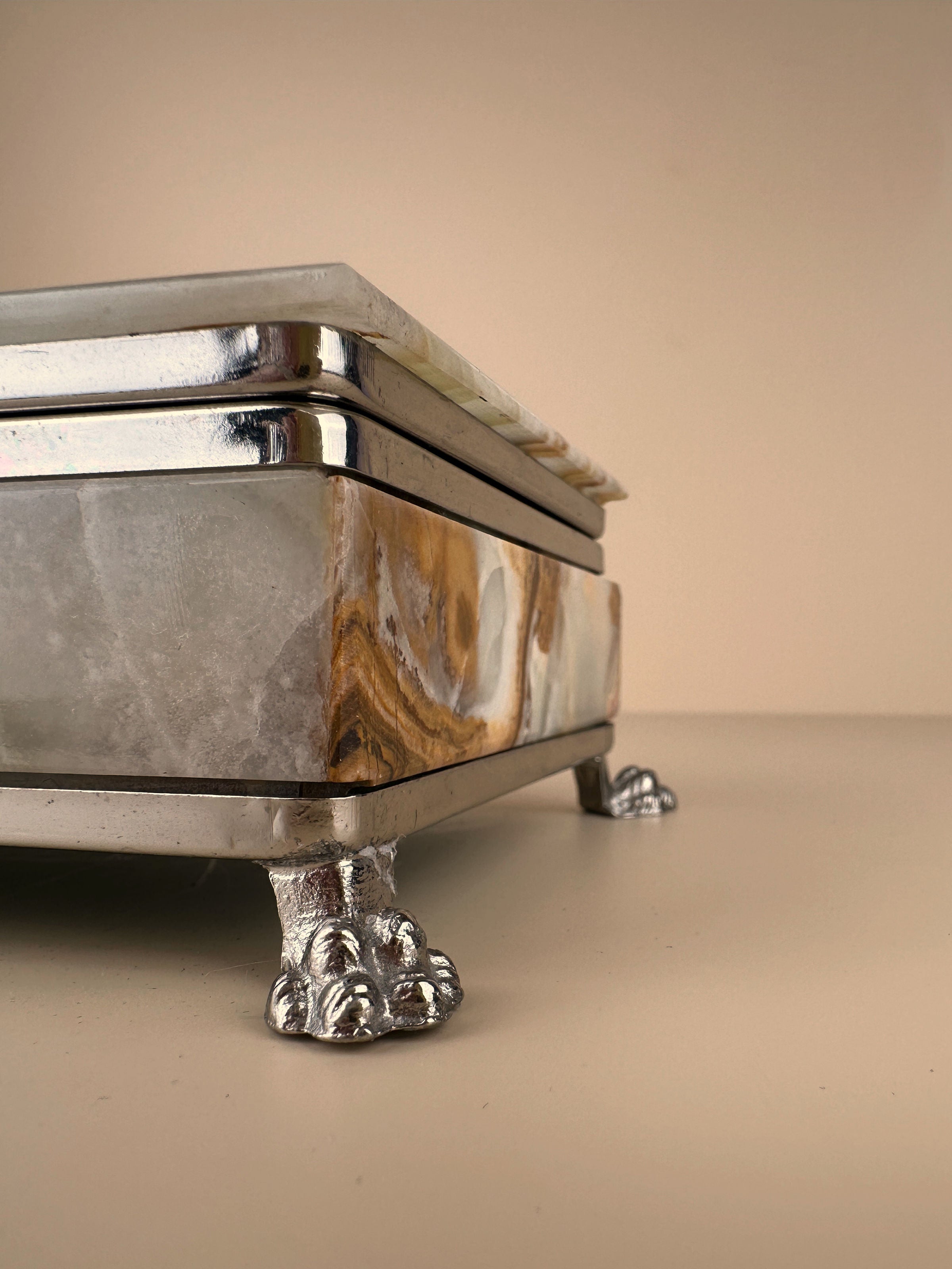 A close-up view of an elegant rectangular vintage Dodo Vintage 1970s Italian onyx jewellery box with metallic trim, resting on ornate silver-colored lion paw feet, against a neutral background. The polished surface of the beige marble showcases a natural pattern of brown, beige, and grey hues.