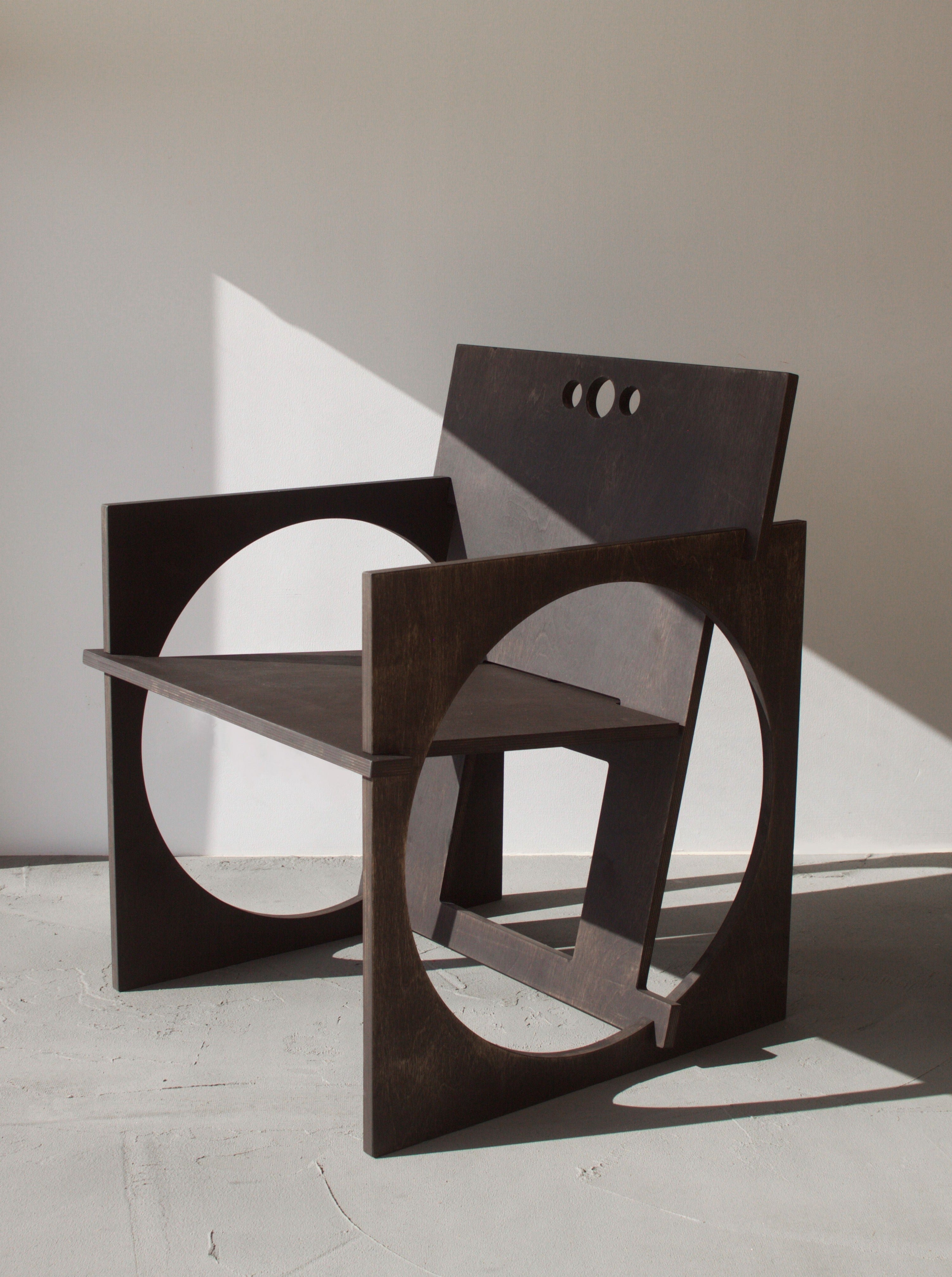 Abstract wooden sculptures featuring geometric shapes and cut-outs, including a Edoardo Lietti Studio Sedia Tonda, displayed in a bright room with sunlight casting shadows on the wall.