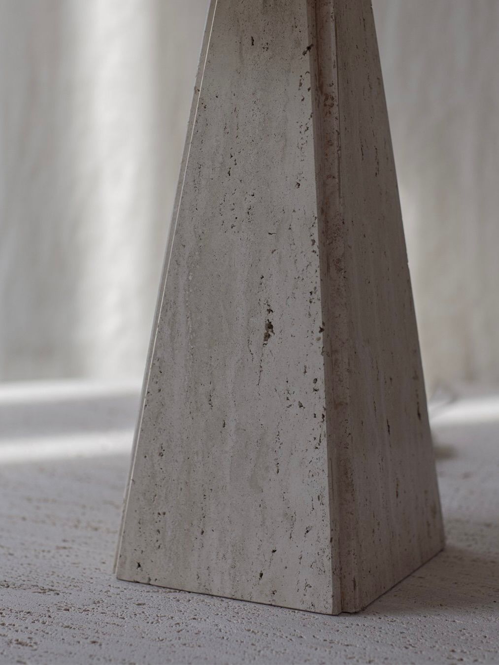 A close-up of a textured, light gray stone pyramid on a rough surface, with a softly blurred background emphasizing its vertical lines and color variations, resembling the elegant design of the Bicci de' Medici Camille Breesch - Travertine Table Lamp.