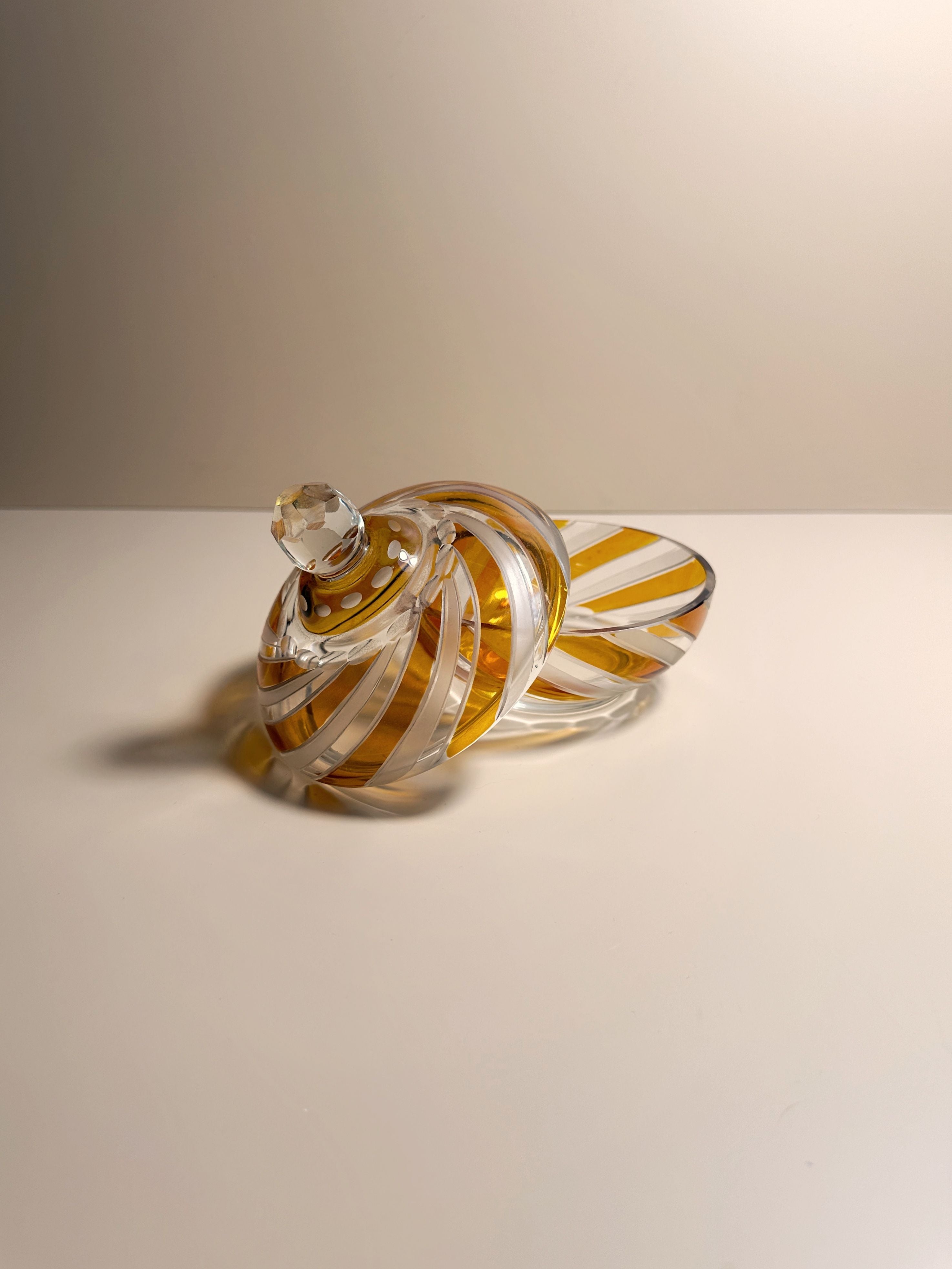 A translucent 1970s Hand-Painted Crystal Bonbonnière by Dodo Vintage, adorned with white and yellow spiral patterns, rests on a smooth, white surface. The image features a softly lit, neutral background that gently highlights the intricate design and craftsmanship of the crystal glasswork.