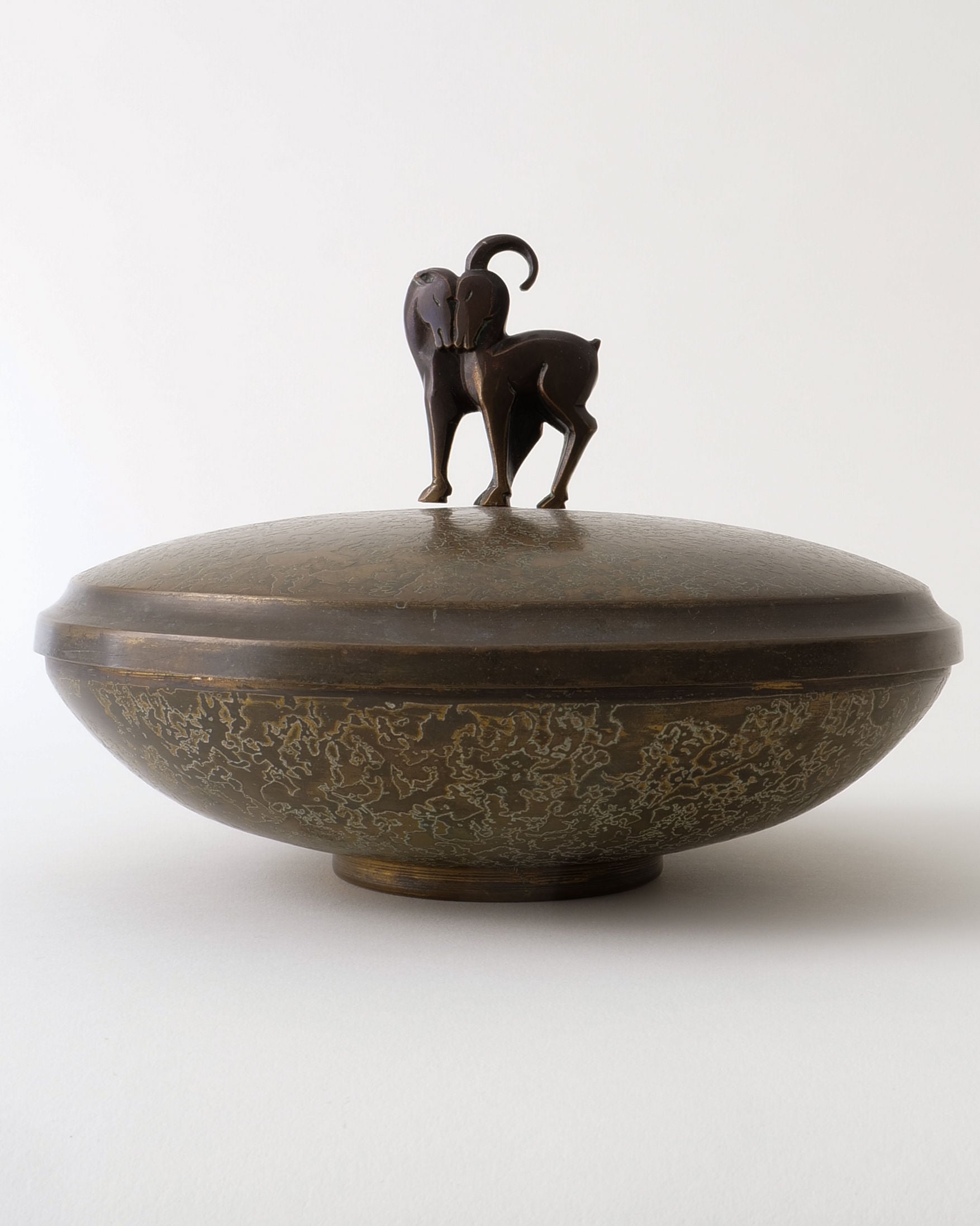 The Deco Box by Pierre Amédée Plasait from Collection apart is a round, bronze Art Deco brass box with a sculpted standing goat lid, featuring curved horns and a mottled textured pattern, shown against a plain white background.