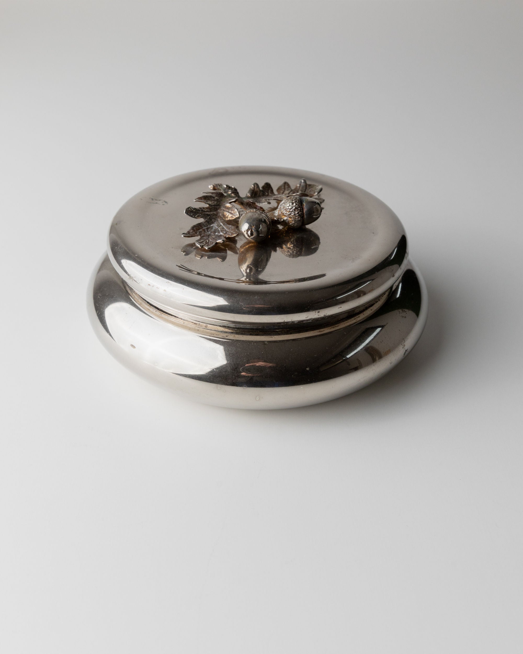 Bottega Jacobs' Silver-plated Box from Italy (1950s) features a shiny, reflective round design. Its lid showcases elegant pinecone and leaf decor akin to vintage tins, complete with an acorn handle for a whimsical touch against a plain white backdrop.