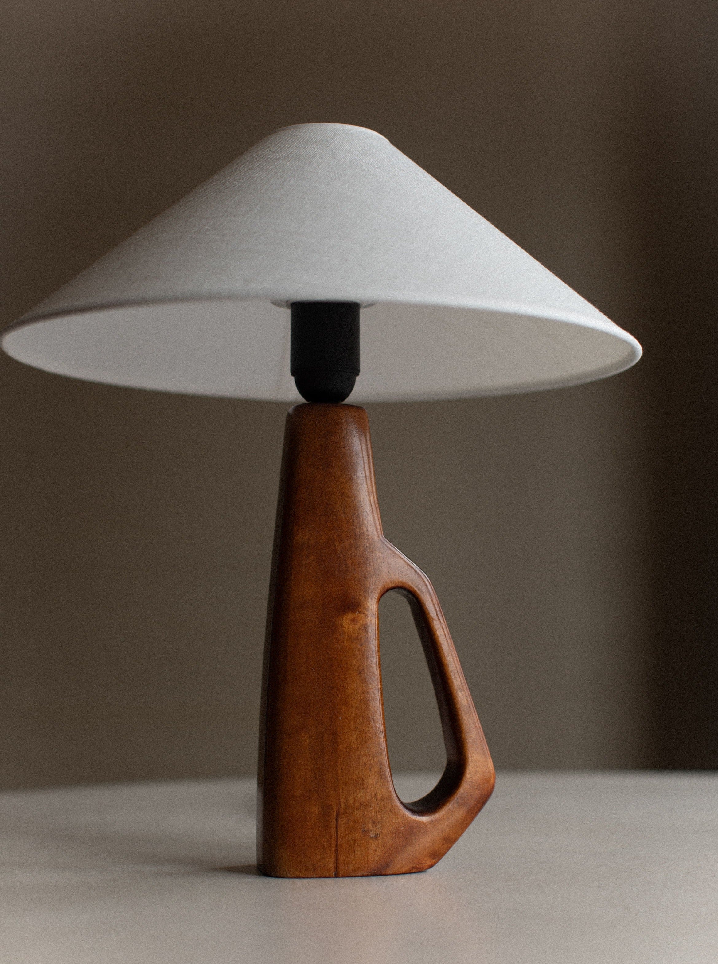 A modern 1960's Wooden Lamp by Out For Lunch featuring a large, white, cone-shaped shade and a distinctive wooden base with hand-carved details and a cutout handle design. The sleek and minimalist aesthetic stands out against a neutral, blurry background.