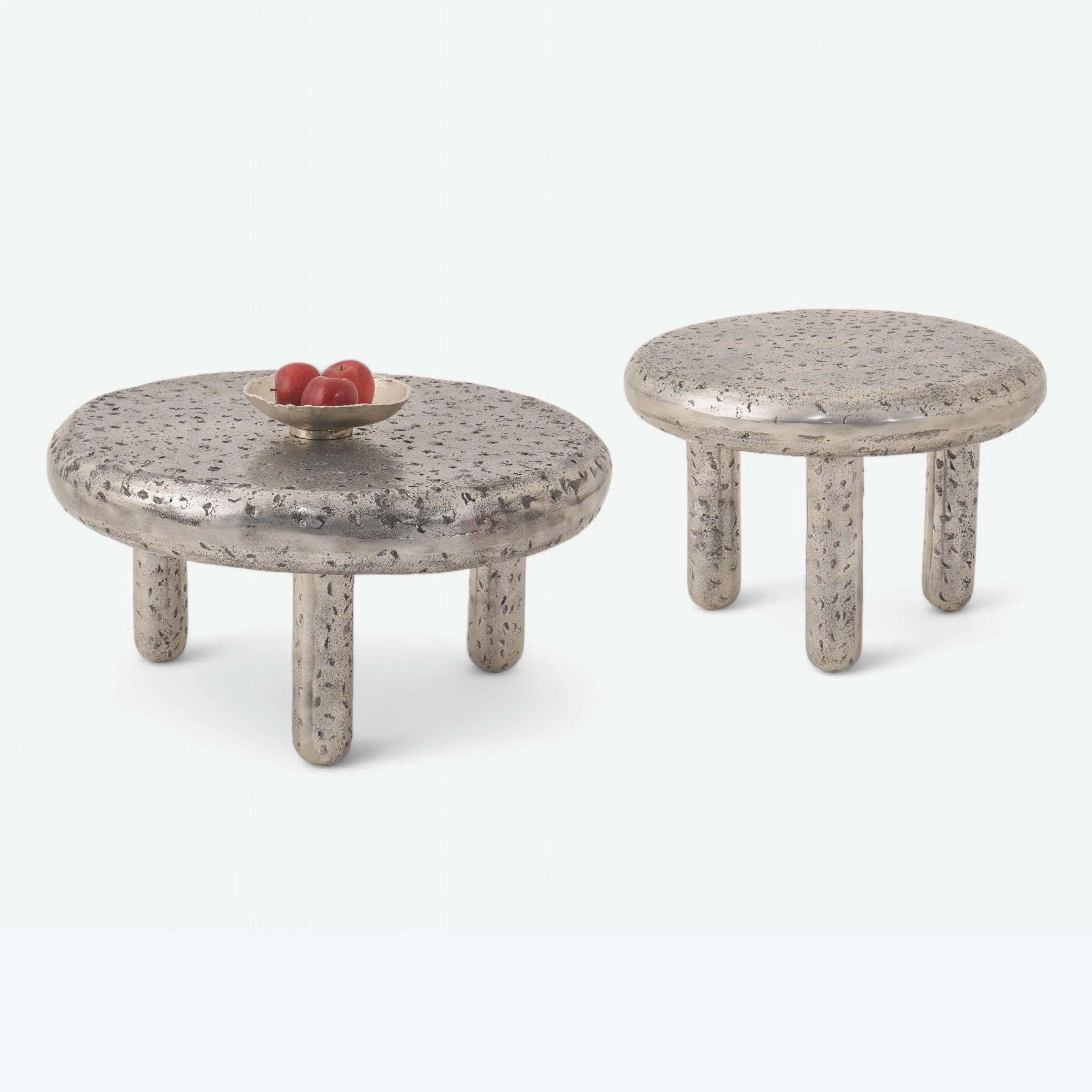 The Crater - Set of 2 by LMNOH features two round metallic tables with textured surfaces and three cylindrical legs, resembling a lunar landscape. The larger table's small wooden bowl holds three red apples, crafting a celestial centerpiece against a modern interior’s plain, light backdrop.