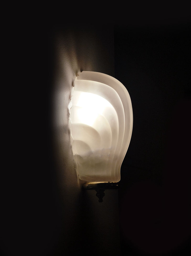 The Les Objoies Shell Wall Light features frosted glass with curved, layered ridges, softly emitting white light and partially illuminating the surrounding wall.