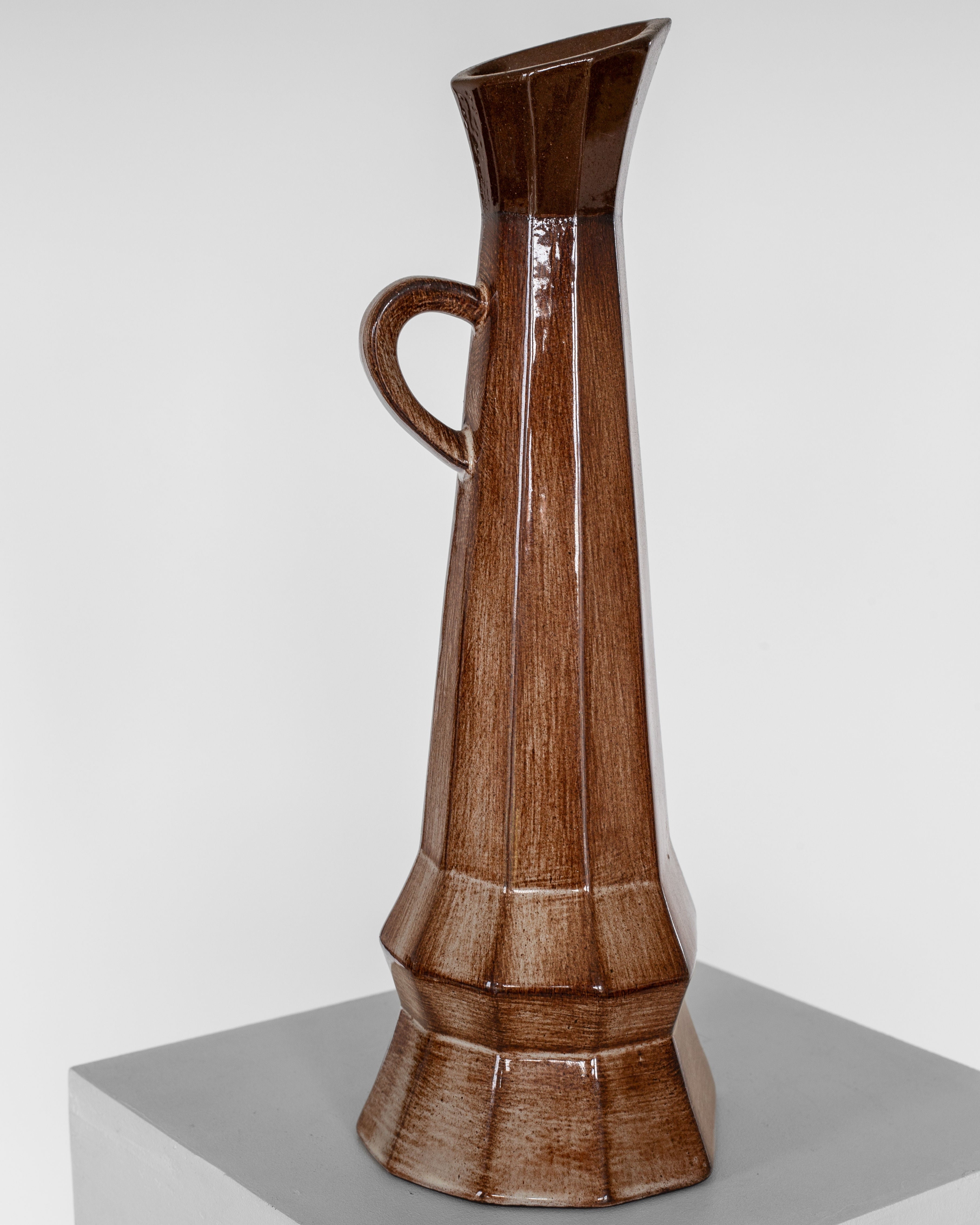 On a gray surface stands the Spigel Cubist Ceramic Pitcher, a tall and glossy brown piece. This uniquely shaped pitcher, evocative of mid-20th-century vase elegance, features a narrow neck, single handle, and geometric facets along its body that lend it an asymmetrical and modern design. The background is plain white.