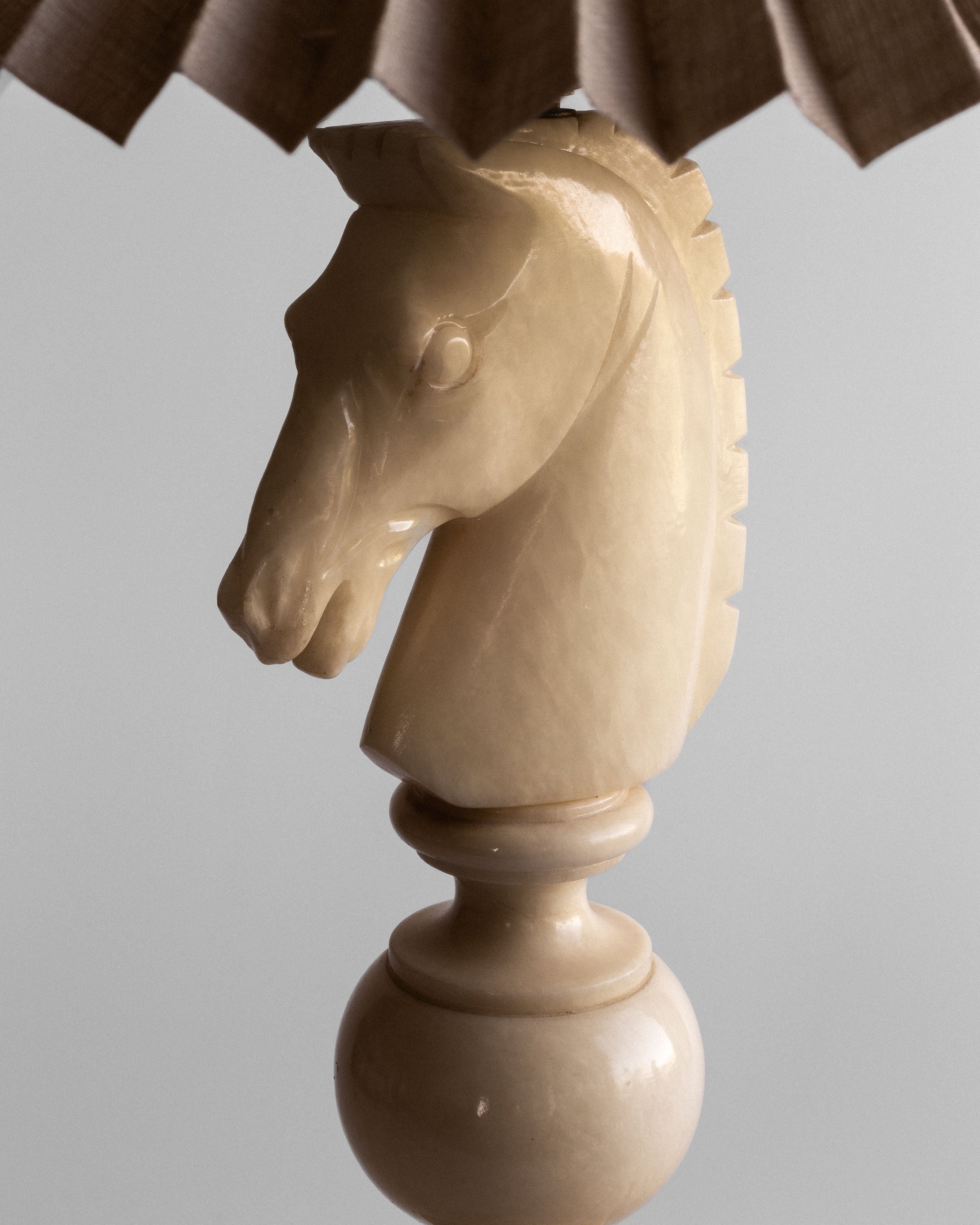 A close-up image of Spigel's White Knight Alabaster Table Lamp from the 1970s, showcasing a sculpture resembling a horse's head made of beige, glossy alabaster. The sculpture is mounted on a spherical base with part of the lamp's pleated vintage Italian lampshade visible at the top against a plain gray background.