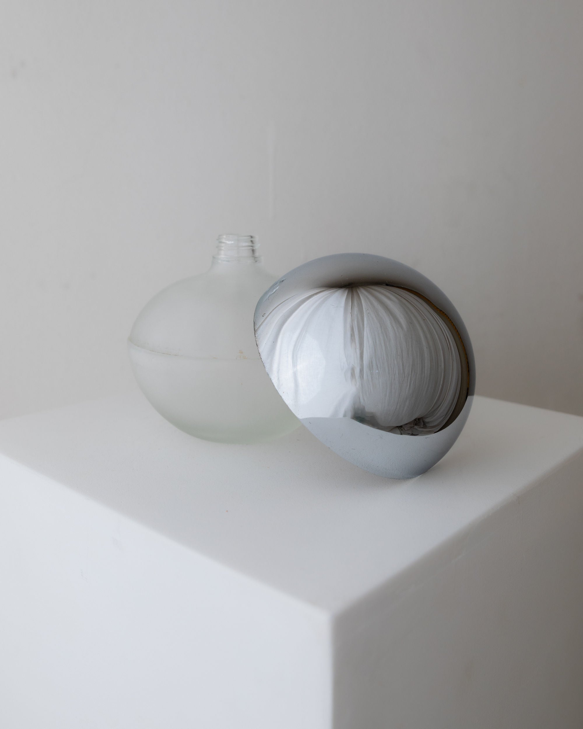 A frosted glass Liqueur Bottle, Italy 1980s by Bottega Jacobs, with an open top and space age design stands on a white pedestal containing a textured, white material. A metallic lid leans against it, reflecting light. The plain light gray backdrop enhances the glass and chrome aesthetic.