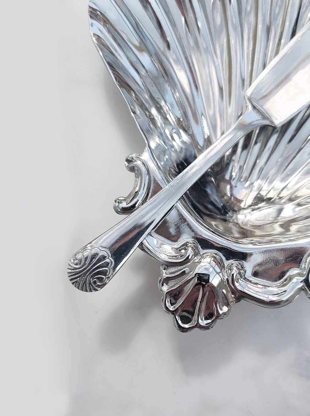 A close-up of the elegant Les Objoies Shell Butter Dish, crafted from stainless steel and featuring a decorative handle, is accompanied by the tip of an assorted knife resting inside. Both the dish and utensil are adorned with ornate patterns that enhance their sophisticated design.