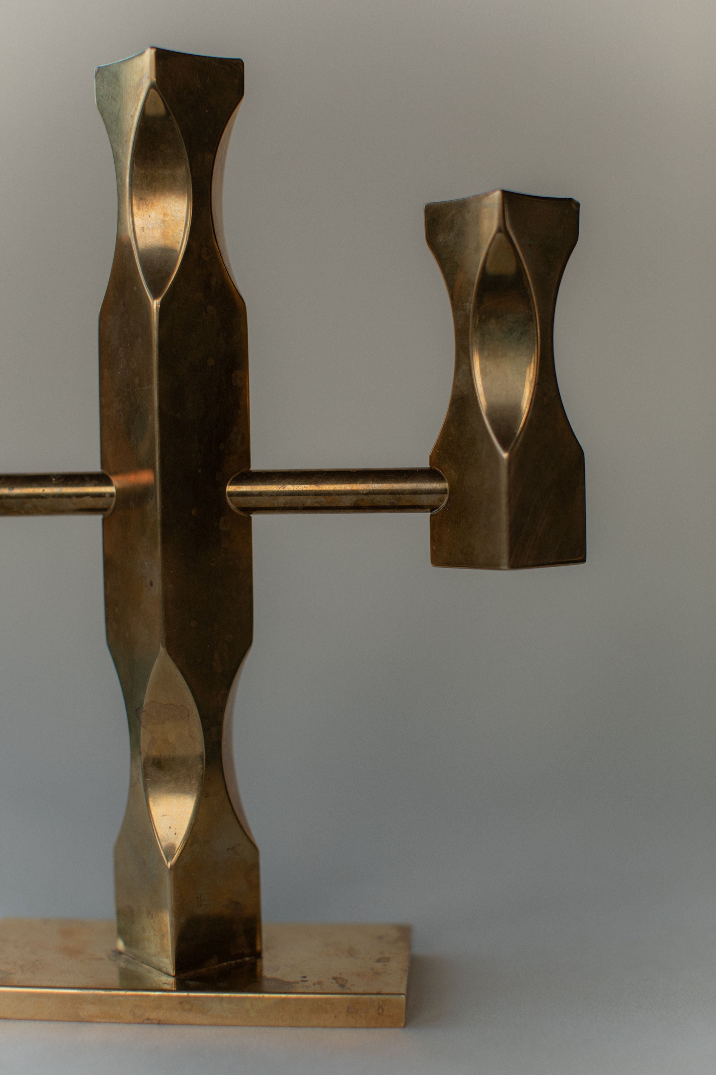 A close-up of the "Brass Candleholder" by Out For Lunch highlights its solid brass, angular design and shiny finish against a neutral backdrop. The piece features vertical and horizontal elements, with a rectangular base resembling a Brutalist candleholder sculpture.