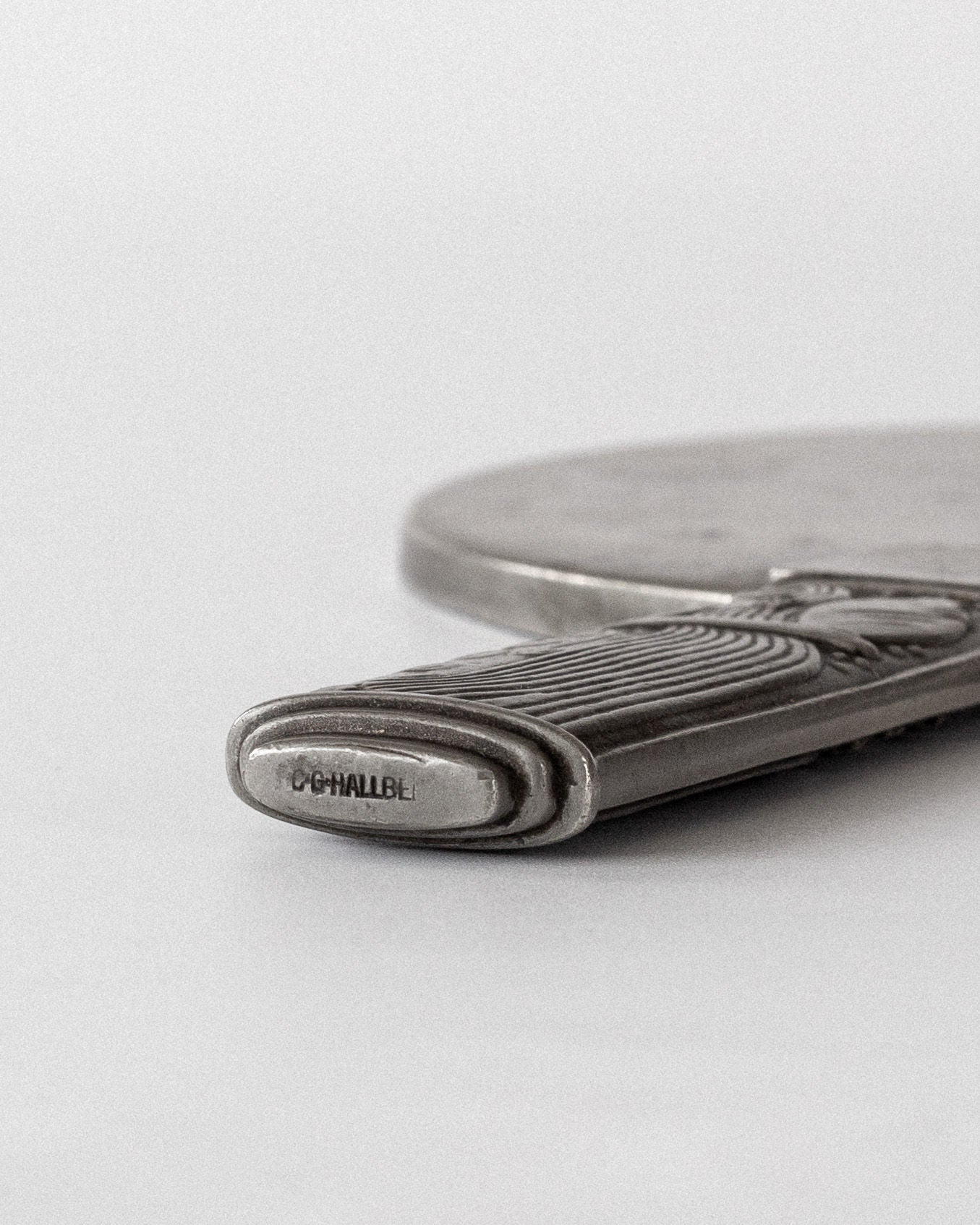 Close-up of a Modernist Metal Mirror by Sylvia Stave from the 1930s, engraved with "O'CHALLBE." The focus is on the handle featuring a modern design from brand Spigel, as the rest softly blurs into the background.