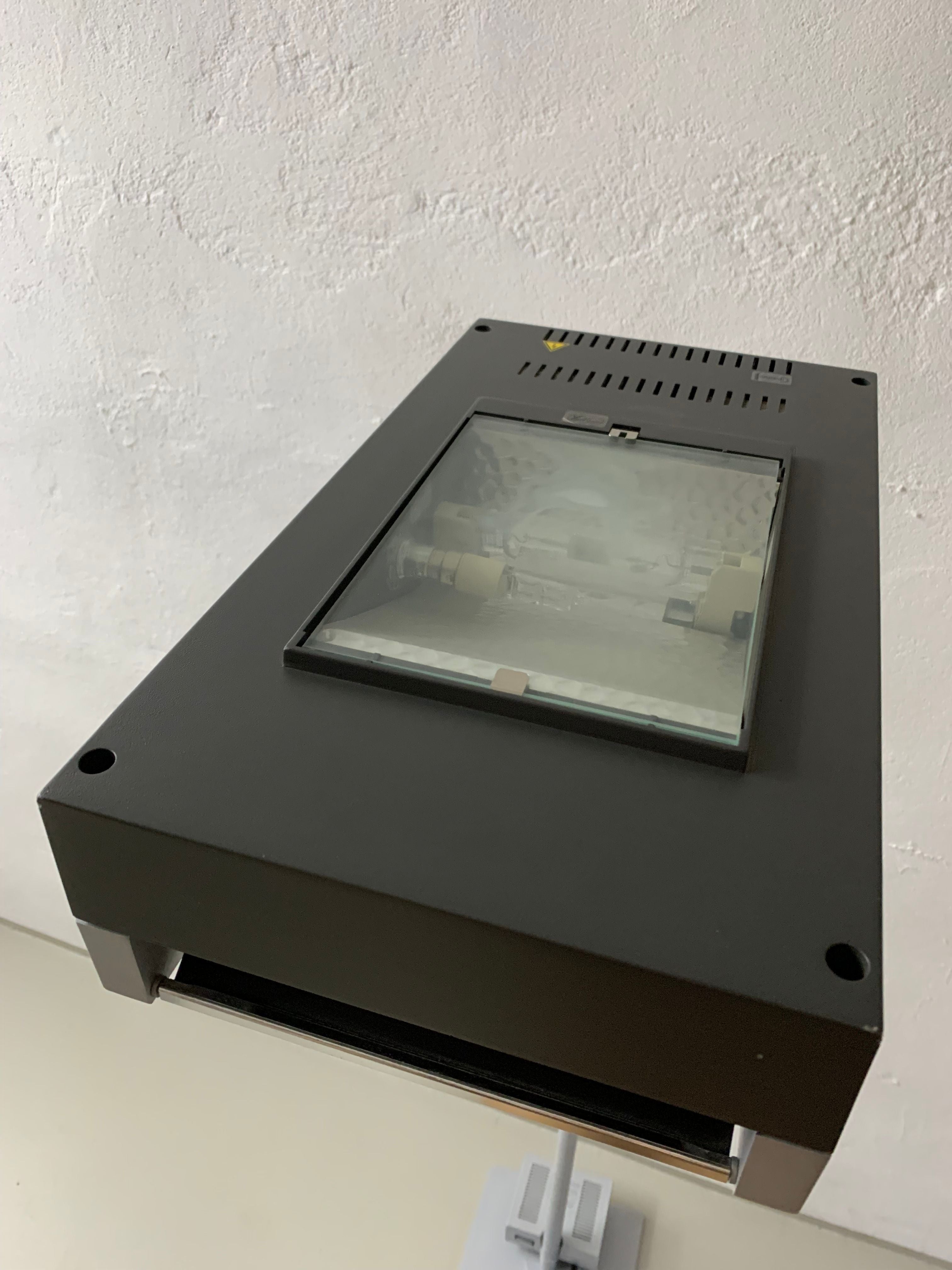 A black slide projector featuring a Veter Vintage Postmodern Metal Lamp by Glen Oliver Löw and Antonio Citterio for Ansorg | 1990s office system, positioned against a white textured wall, showing a focused image of a car within its display screen.