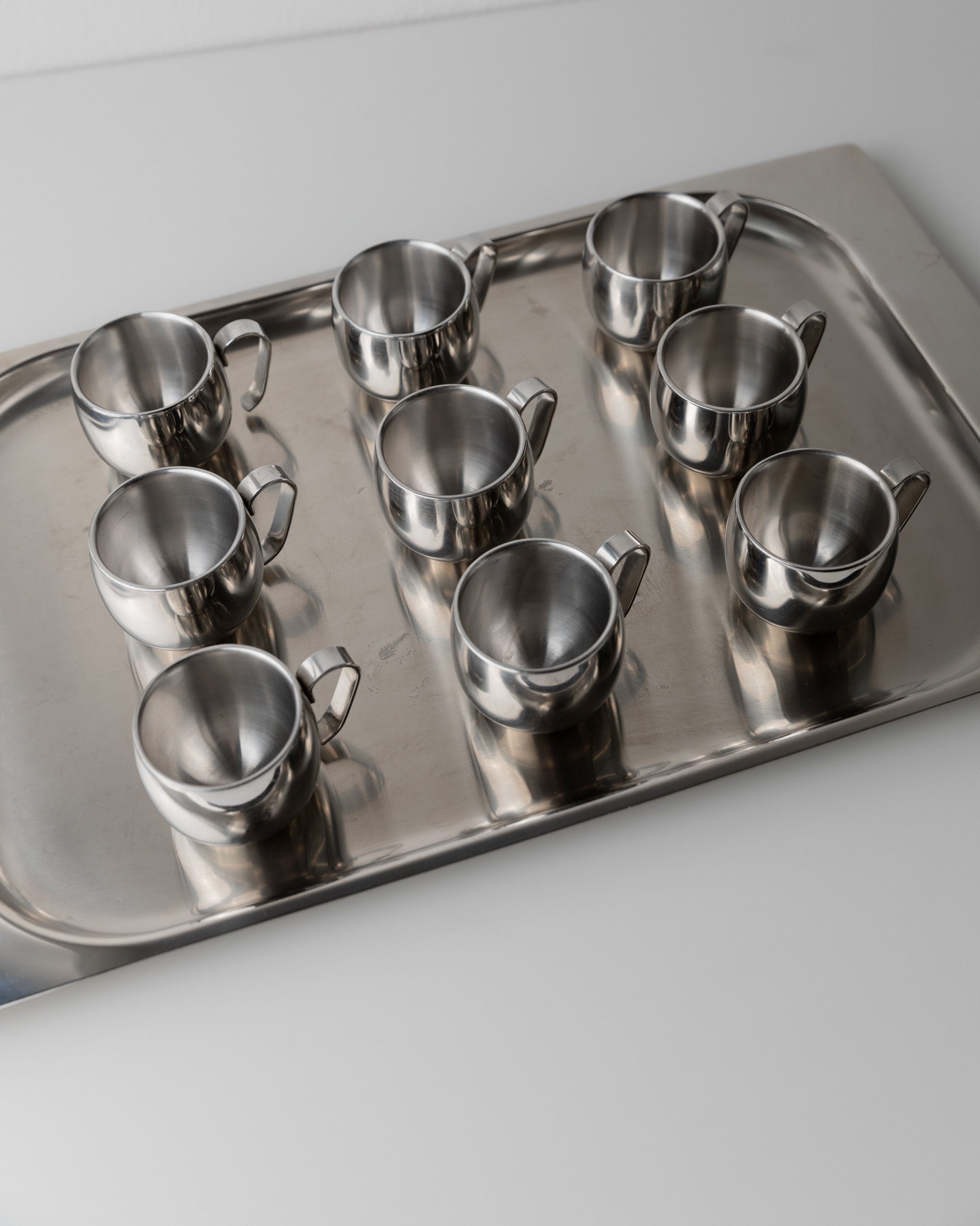 The Coffee set by IMCO 70s from Bottega Jacobs presents eight sleek stainless steel cups on a metal tray, arranged in two tidy rows of four, embodying minimalist design on a light surface.