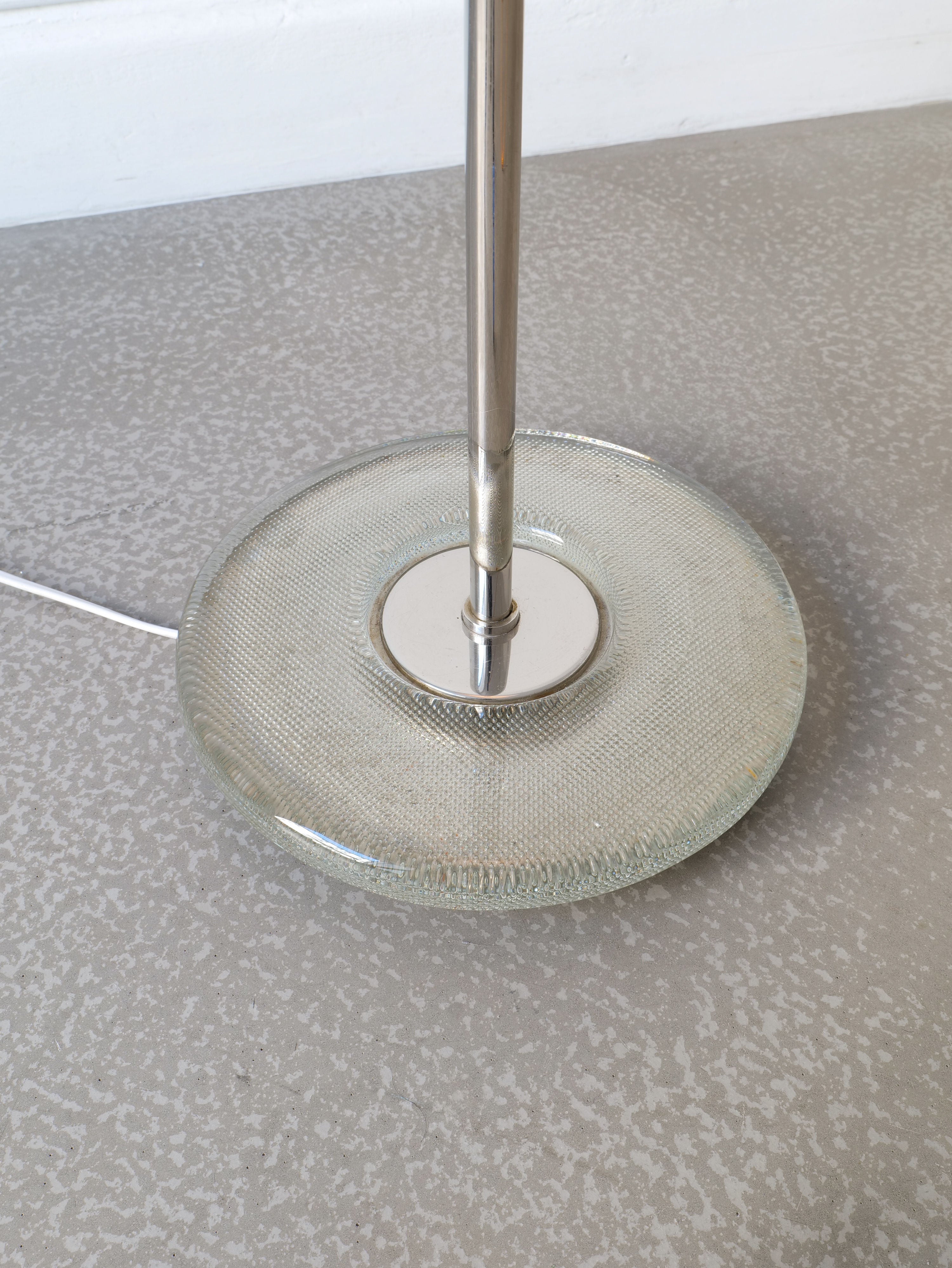 A close-up of a Deco Floor Lamp Harald Notini 1940s by Collection apart base featuring a textured, circular glass disc with a metallic rod extending vertically from its center. The lamp stands on a speckled gray surface, and the white power cord is slightly visible on the left side.