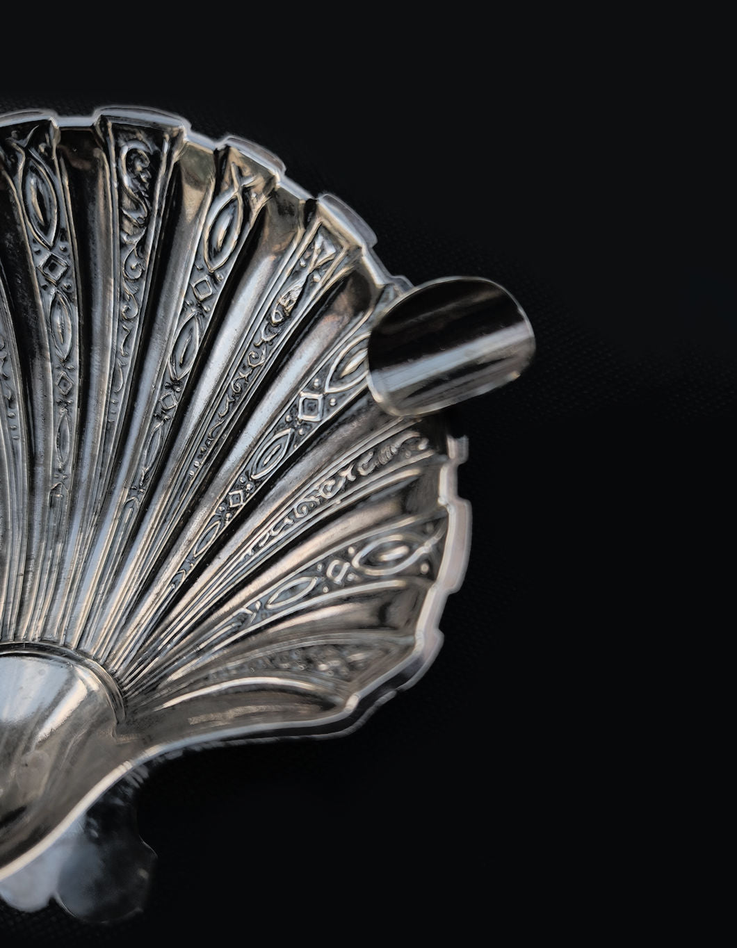 A close-up of an ornate Shell Ashtray by Les Objoies in a silver hue, showcasing intricate, decorative engravings reminiscent of a shell against a dark background. The spout-like detail is visible on the right side.