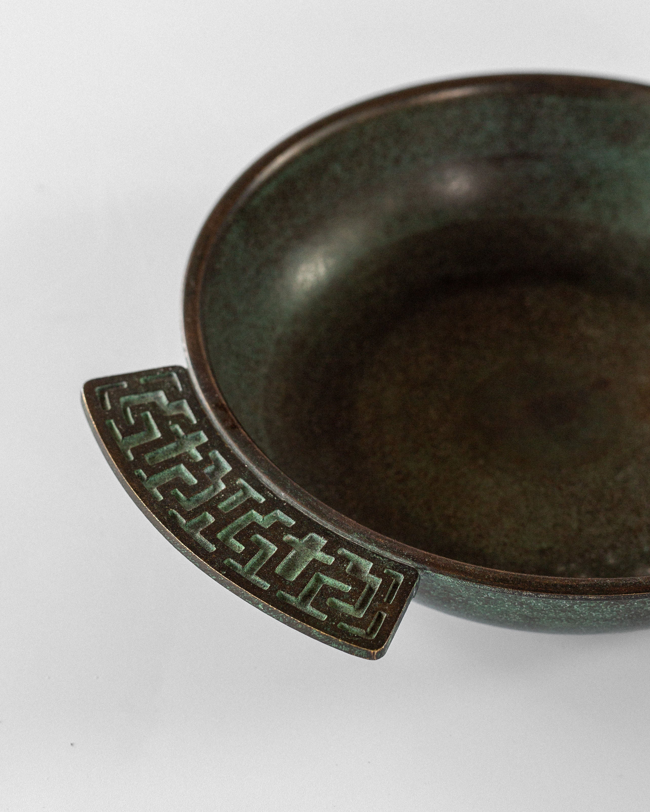 The Spigel Art Deco Bronze Bowl from Sweden, 1930s, features a round ceramic design with a bronze color and dark patina over a light background. It has Greek key handles and a slightly speckled texture that evokes rustic charm.