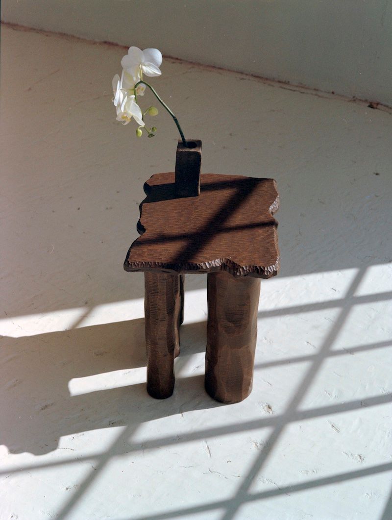 The Mare Side Table by Ombia Studio, a small rustic piece crafted from solid white oak with uneven edges, stands on a sunlit floor. It has three legs, supporting a single white orchid in a slender vase, with window pane shadows enhancing its natural beauty.