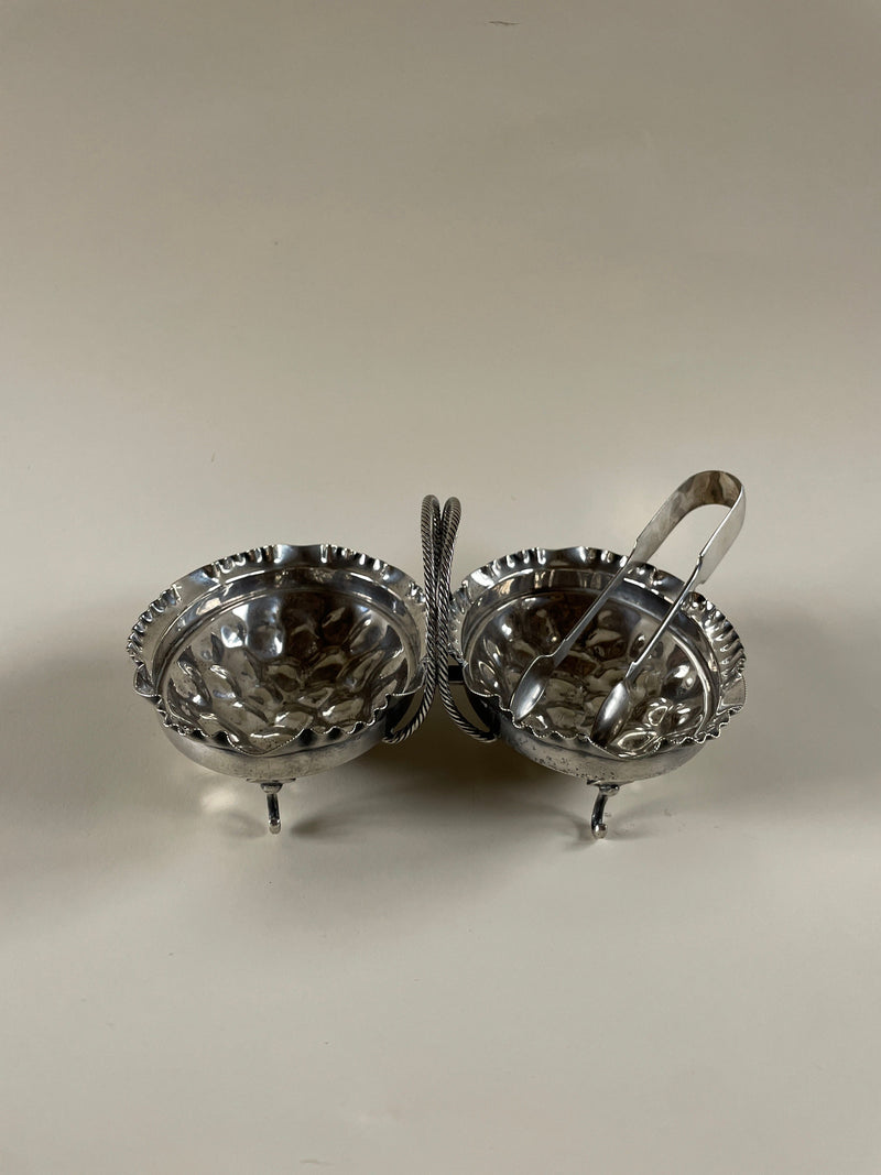 Introducing the Maud Vaughan Silver Plate Serving Dish, a vintage decorative piece crafted from metal and plated in silver. This exquisite dish showcases two round compartments elevated on small legs, adorned with ornate details. A small set of tongs rests elegantly on one side against a plain light background, enhancing its sophisticated charm.