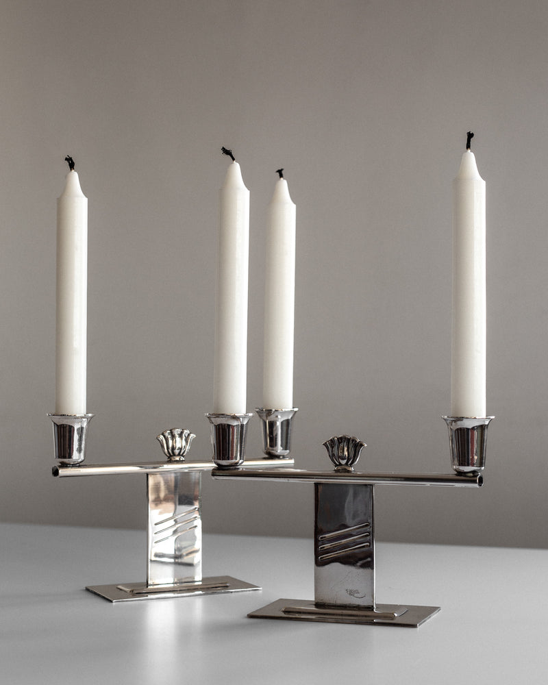 A pair of Spigel Silver-Plated Art Deco Candle Holders, featuring sleek geometric designs, hold unlit white taper candles. They rest on a light gray surface against a plain background. The elegant metalwork harmonizes beautifully with the simplicity of the candles, reflecting Scandinavian craftsmanship.