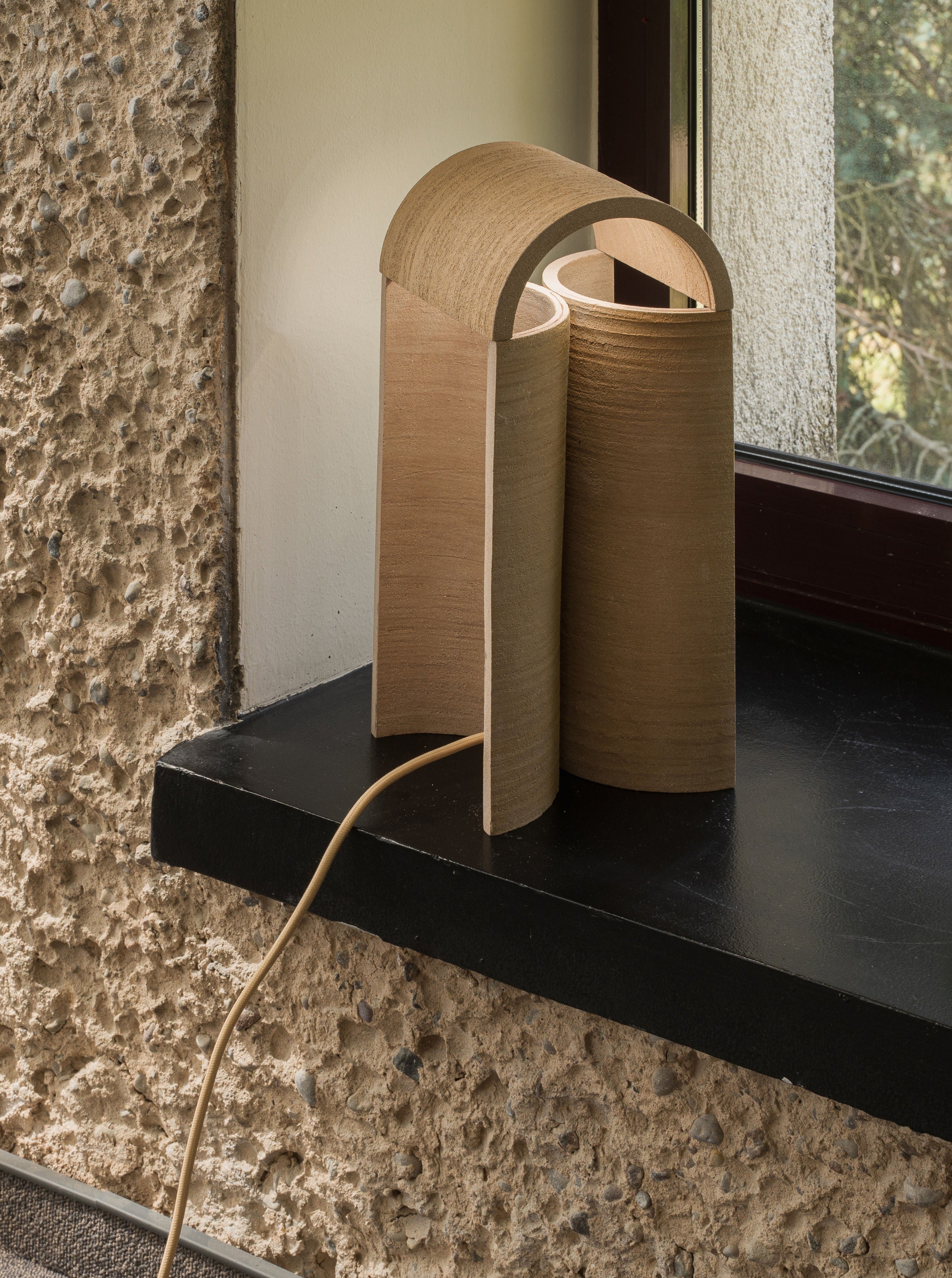 A modern, curved wooden table lamp with an arch design sits on a black windowsill near a textured stone wall. This piece of sculptural lighting, the "Layers of..." Light II by Evelina Kudabaite, is plugged into an outlet with a visible cord extending from its base. Sunlight filters through the window, softly illuminating the scene.