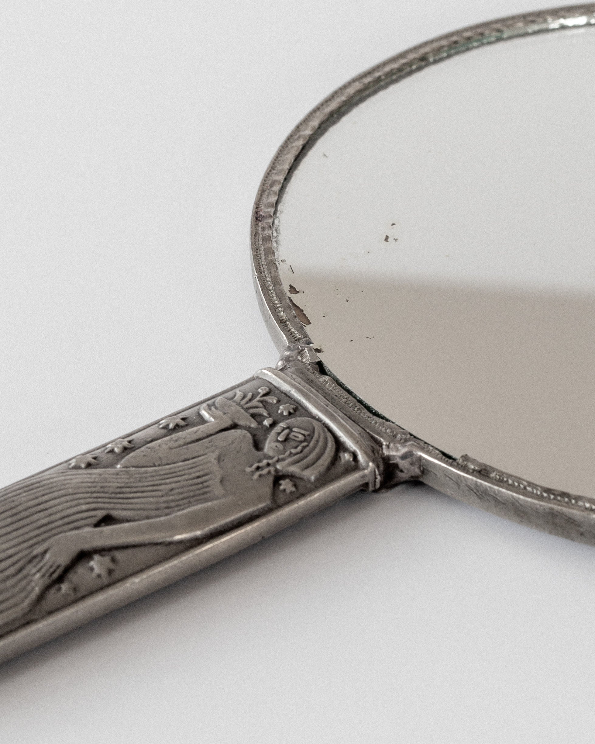 The Modernist Metal Mirror by Sylvia Stave from Spigel, crafted in the 1930s, features a pewter handle and border with an engraved figure and star pattern. Showing signs of wear and some tarnish, it evokes classic craftsmanship on a simple grey background.