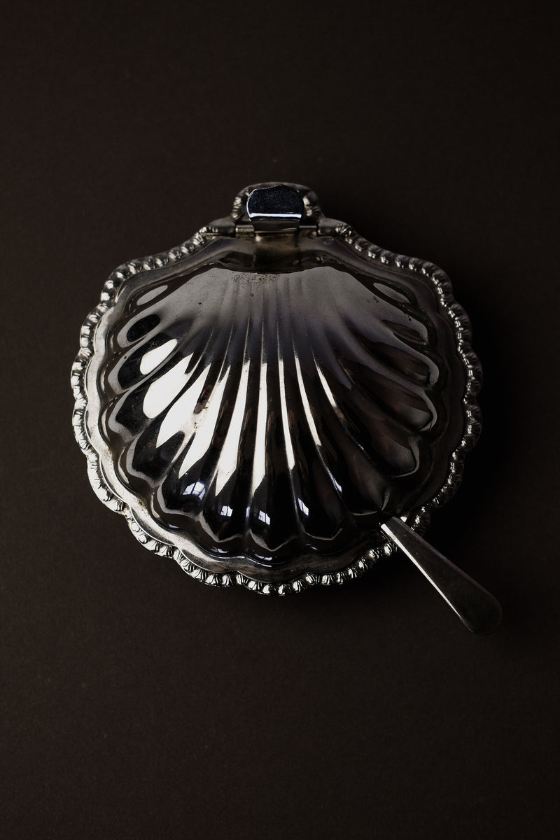 The septembre studios Shell Shaped Butter Dish with Spoon boasts a vintage brass seashell design, complete with an attached spoon and clasp. Intricate edge details enhance its elegance, making it a sophisticated addition to any dining setting against a dark background.
