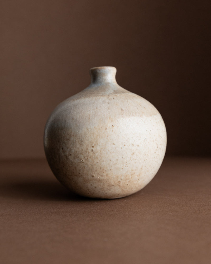 The Ceramic Vase by Bernd Röter (1970s) from Bottega Jacobs is small, round, vintage-style with a narrow neck and textured speckled surface, blending seamlessly into the earthy tones of the brown background.