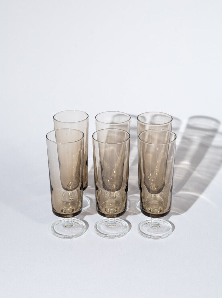 Six Smokey Grey Champagne Glasses by Luminarc from Break The Mould Magasin, with clear bases, reminiscent of vintage champagne glasses, are arranged in two rows on a white surface. The glasses cast soft shadows to the right, indicating light coming from the left. The background is plain, emphasizing the minimalist setting.