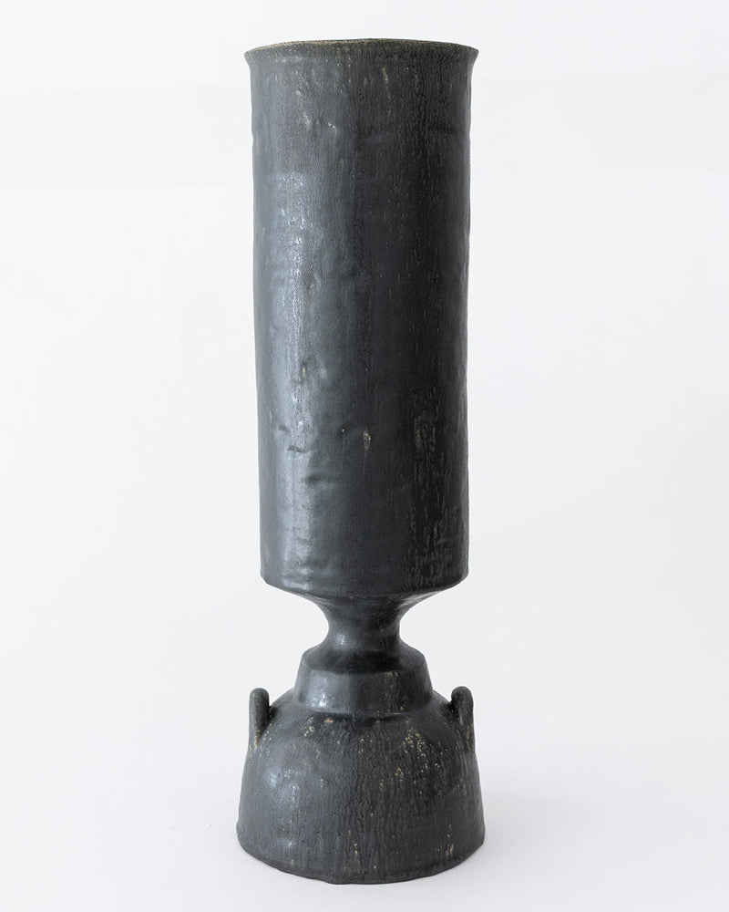 The Isolated n.32 Vase by Canoa Lab is a tall, cylindrical dark ceramic piece with a textured surface and flared rounded bottom, reminiscent of ancient artifacts, displayed on a plain white background.