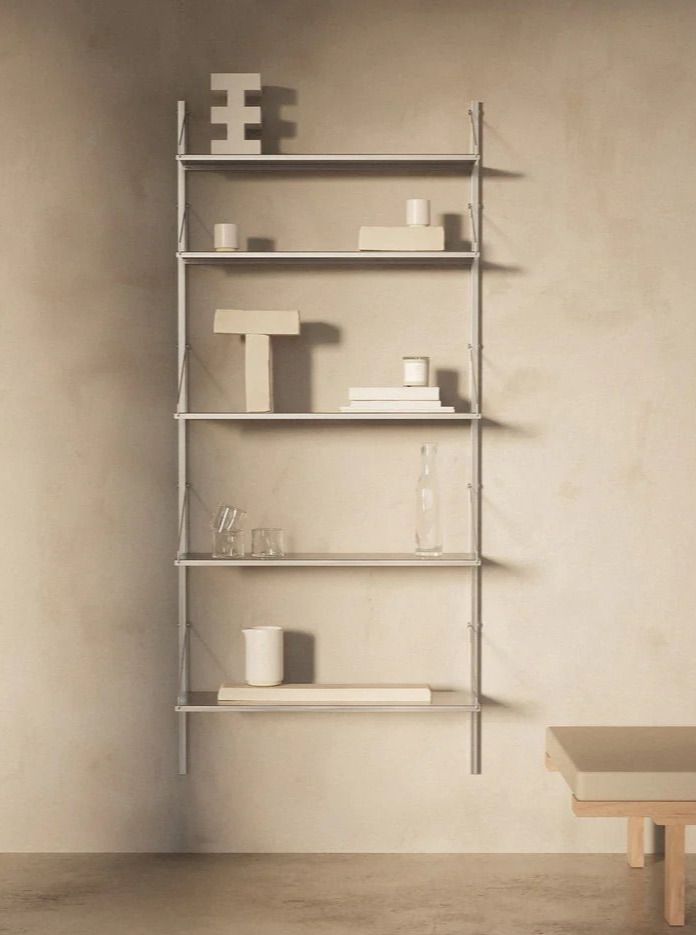 A minimalist FRAMA Shelf Library Single Section | Stainless Steel | H1852 / W80 with five stainless steel shelves holds various geometric decor pieces, including a block-like sculpture, a T-shaped object, books, and vases. A wooden table with a bench is partially visible to the right against a beige wall.