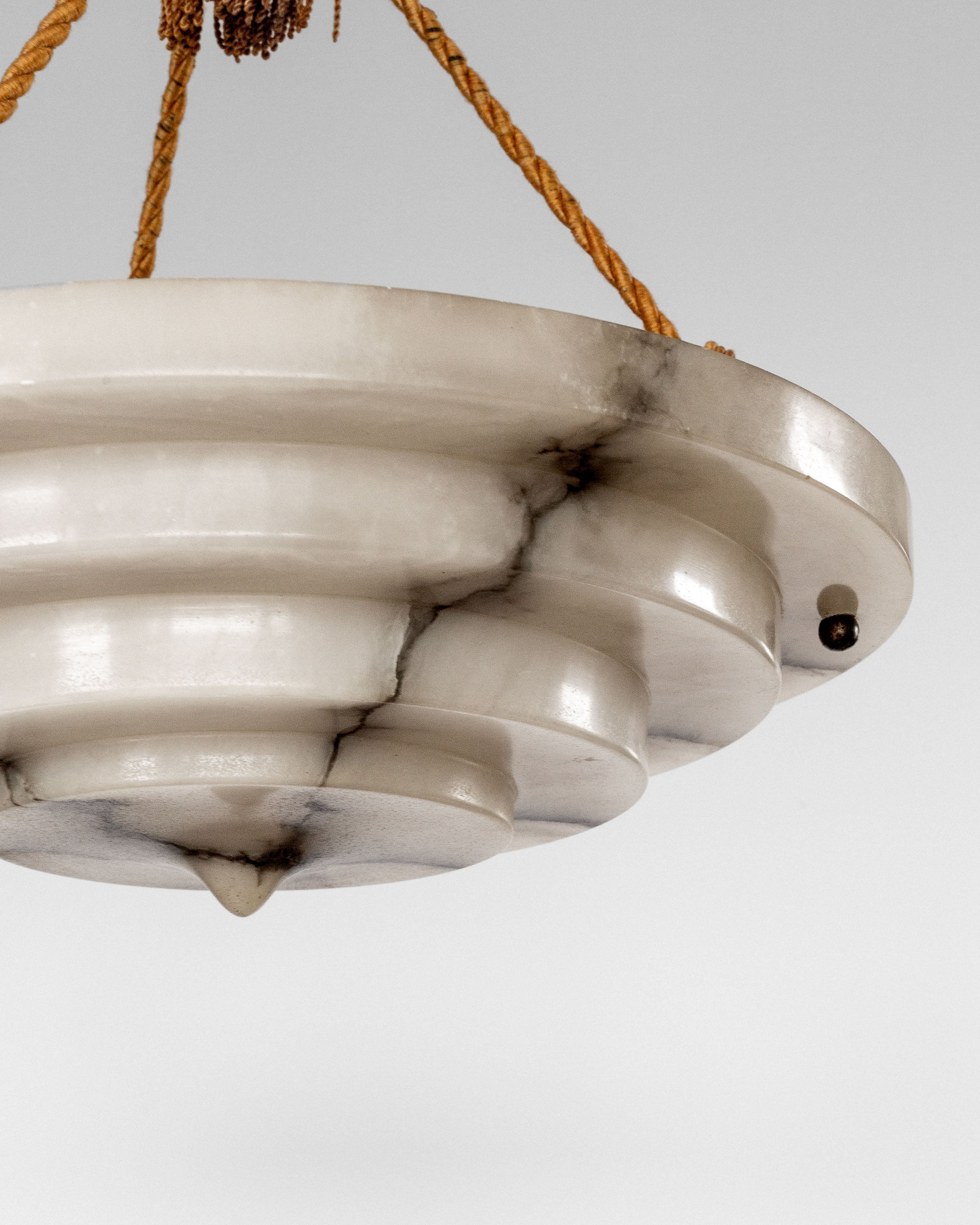 The Spigel Art Deco Alabaster Pendant Chandelier showcases a tiered circular design inspired by Swedish Grace, with a polished marble surface streaked with dark veins. It's suspended by three brown ropes and features an iconic small pointed element at the bottom, exemplifying period lighting.