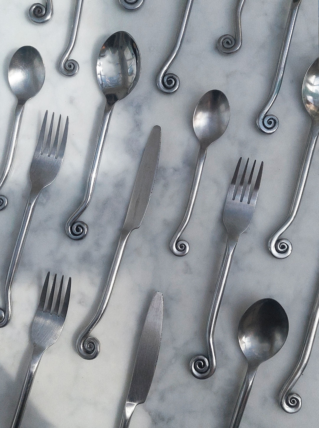 A set of four utensils from Les Objoies, featuring their Swirling Cutlery collection with forks, spoons, and knives spread across a marble backdrop. Each piece is designed with unique curled handles, creating an eye-catching pattern ideal for any sophisticated table setting.