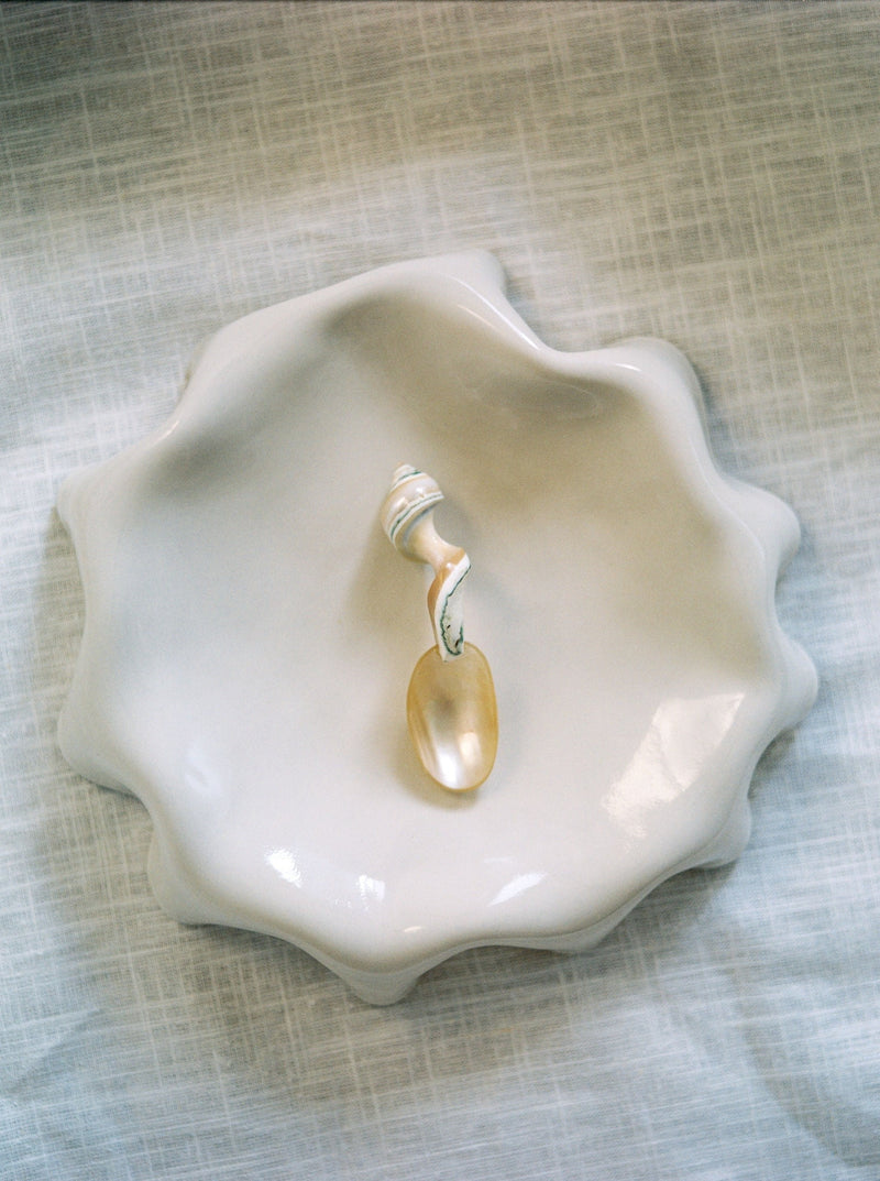 A wavy-edged, glossy Melted Butter Plate in Off White from LESS graces a light, textured surface. Resting within the dish is a small spoon with a handle crafted from a white seashell, displaying hints of beige and gray. This delicate coastal-themed aesthetic exemplifies practical tableware at its finest.