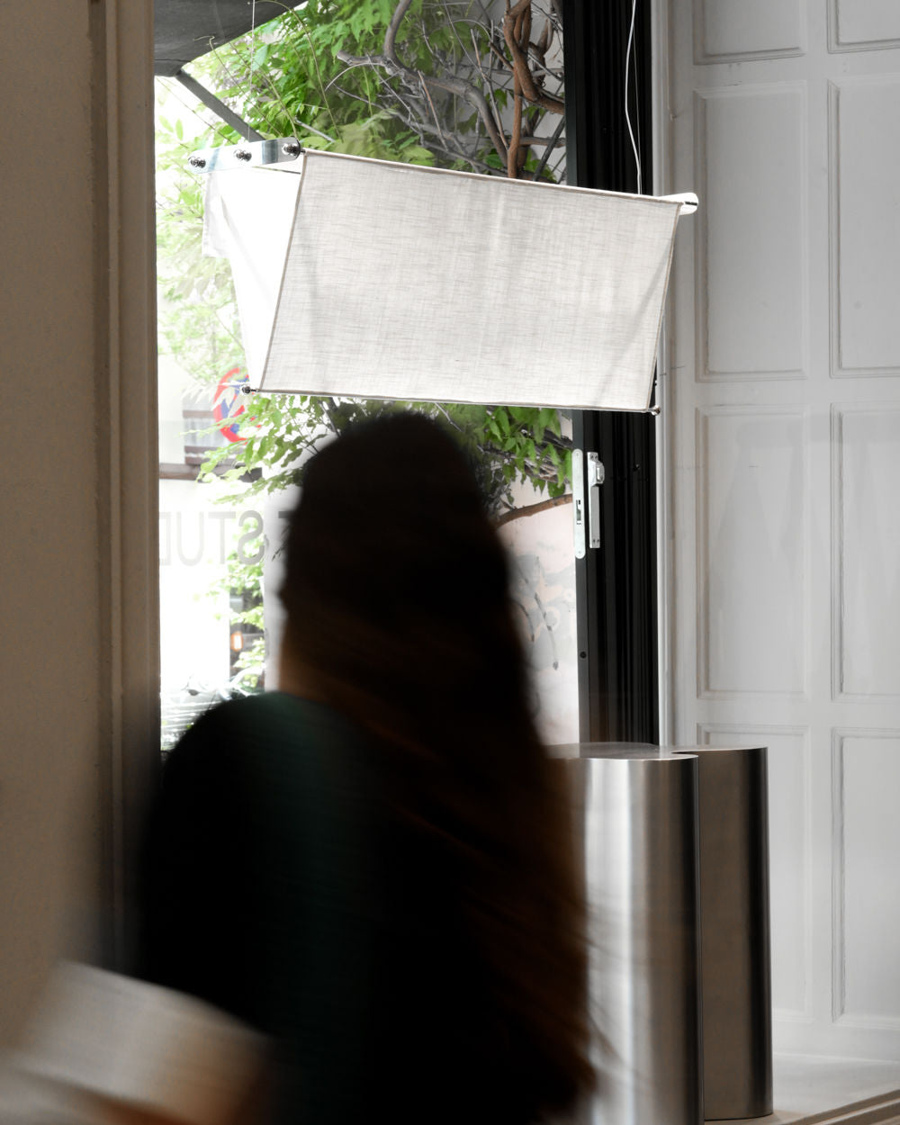 A person with long hair walks indoors near a window with sheer curtains. Minimalist decor features the Caia Leifsdotter Vento Pendant and a stainless steel base, while green leaves and natural light shine outside.