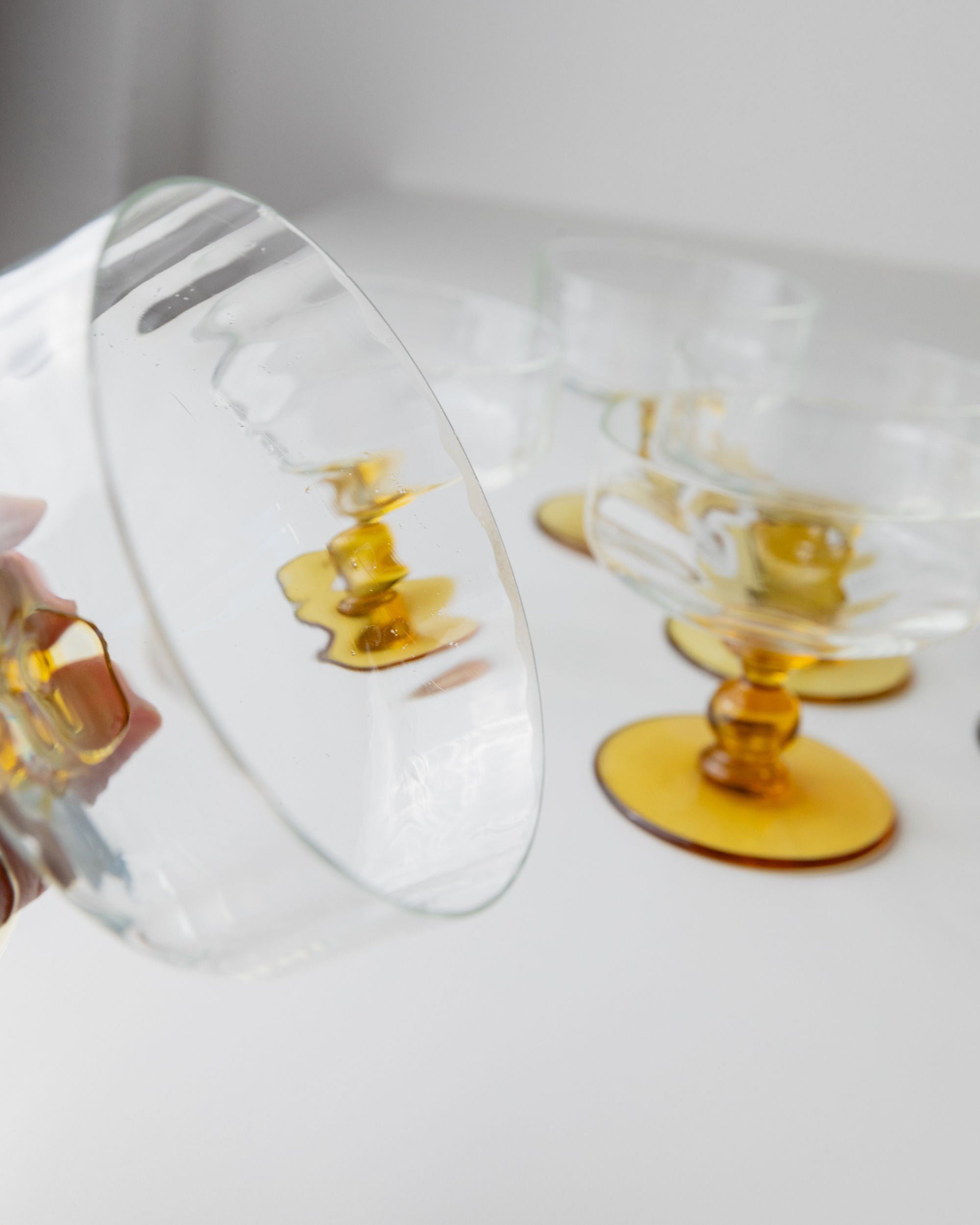 Close-up of a hand holding a clear glass with an amber stem from Bottega Jacobs' Set of 6 Murano Glass, Italy 70s. More similar glasses are visible in the background on a white surface.