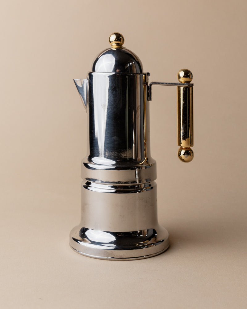 The Coffee Set by VEV Italy 80s from Bottega Jacobs features a shiny, metallic Italian mocha pot with gold handle and knob, standing upright on a beige surface against a beige background—evoking vintage charm.