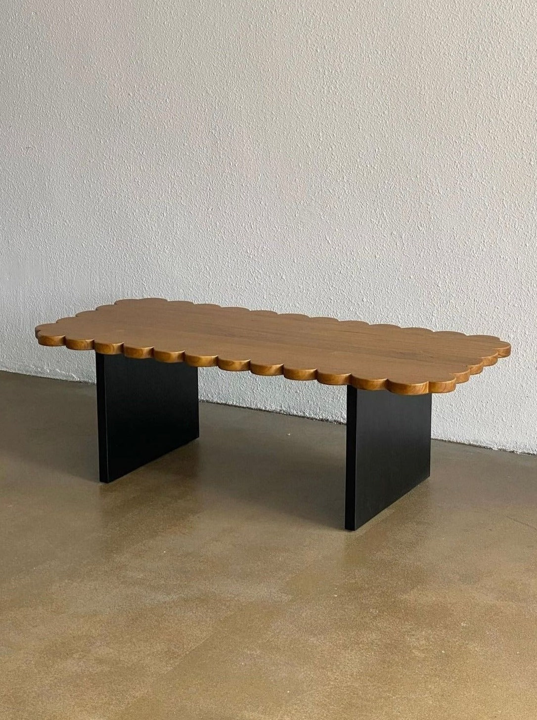 The Biskut Coffee Table by Studio Kallang, with handcrafted scallop detailing on top, supported by two black rectangular legs, stands on a polished concrete floor against a plain white wall.