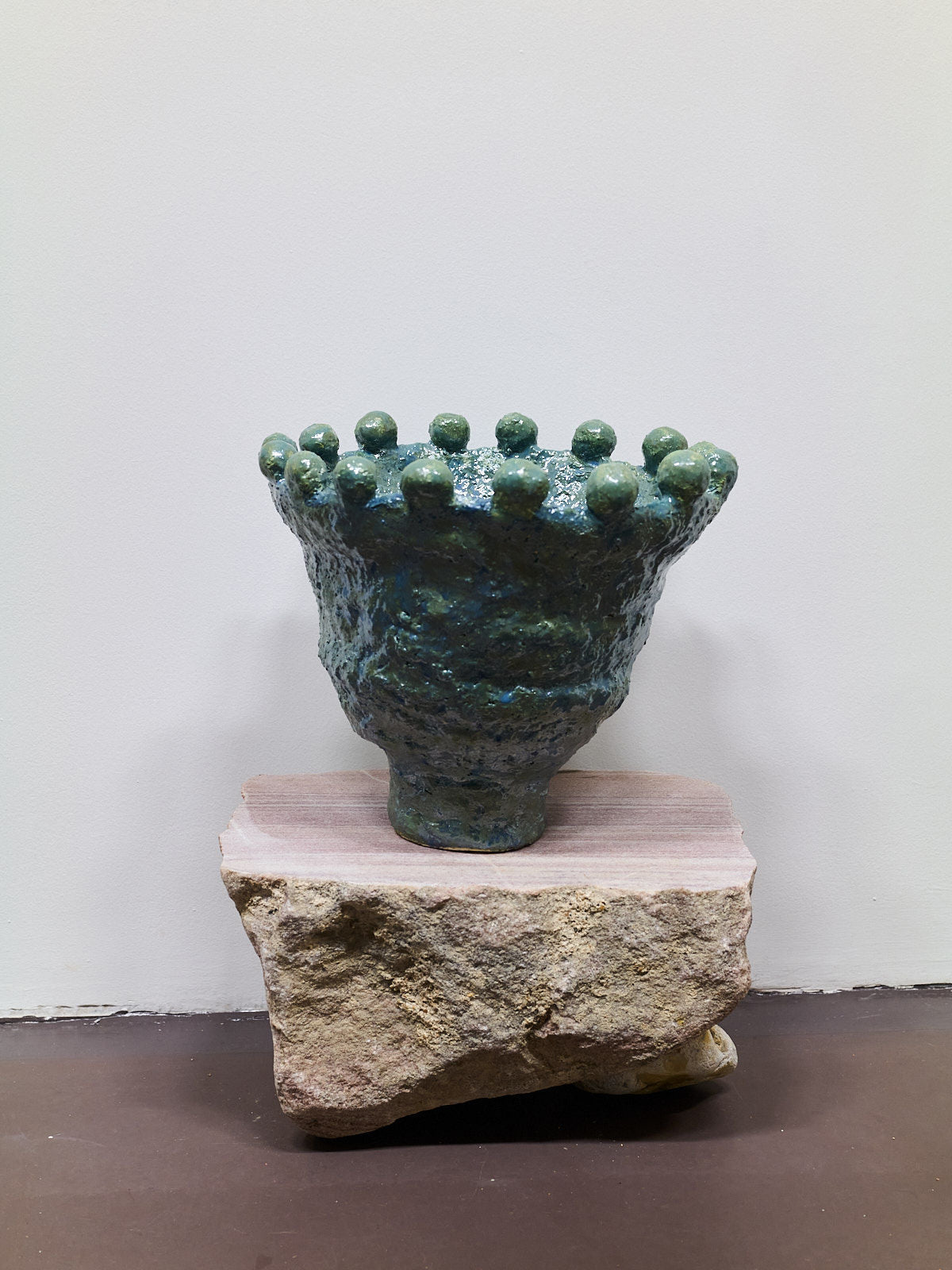 The Stoneware Coupe by Claire Pain is a dark green ceramic vase with a textured design, flared top, and round rim protrusions, finished with metallic enamel. It sits on a beige stone base against a plain, light-colored background.