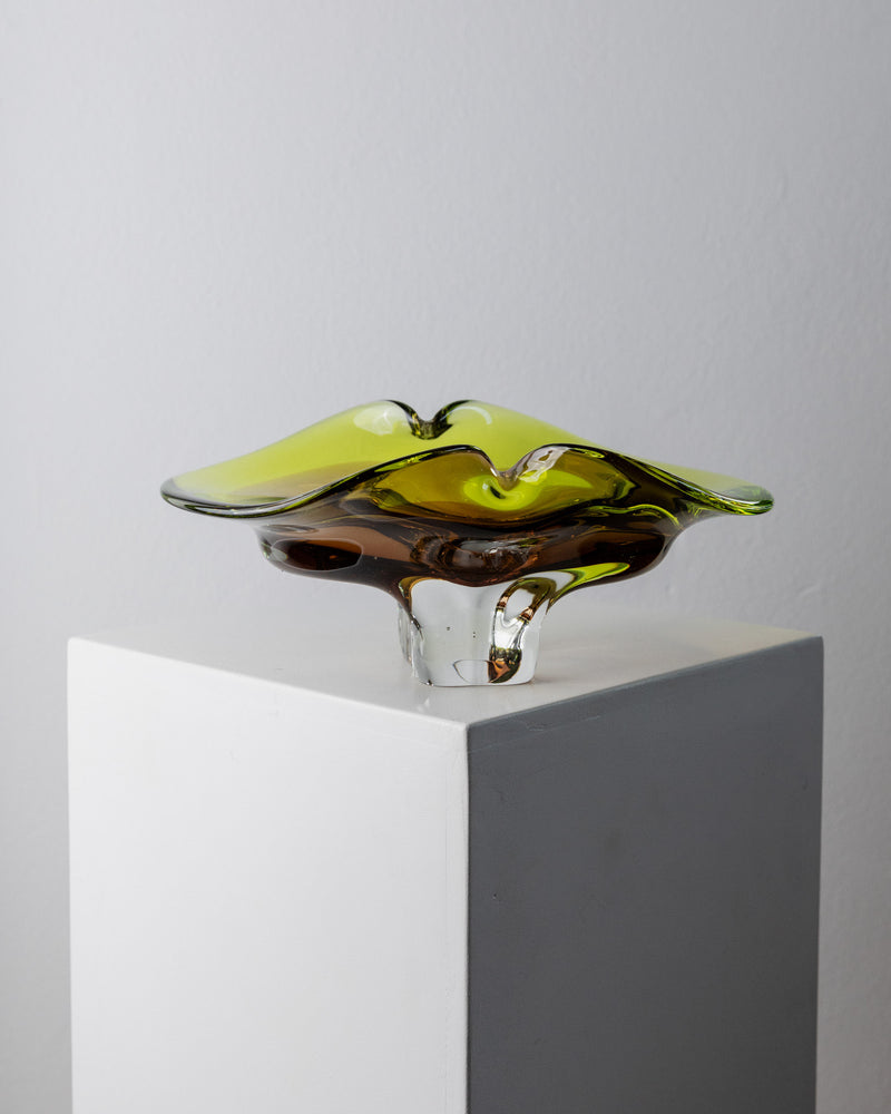 A Murano Glass Bowl from the 1970s by Bottega Jacobs, featuring yellow and clear sculpted glass with wavy edges, sits on a white pedestal, highlighting exquisite Italian craftsmanship against a plain background.