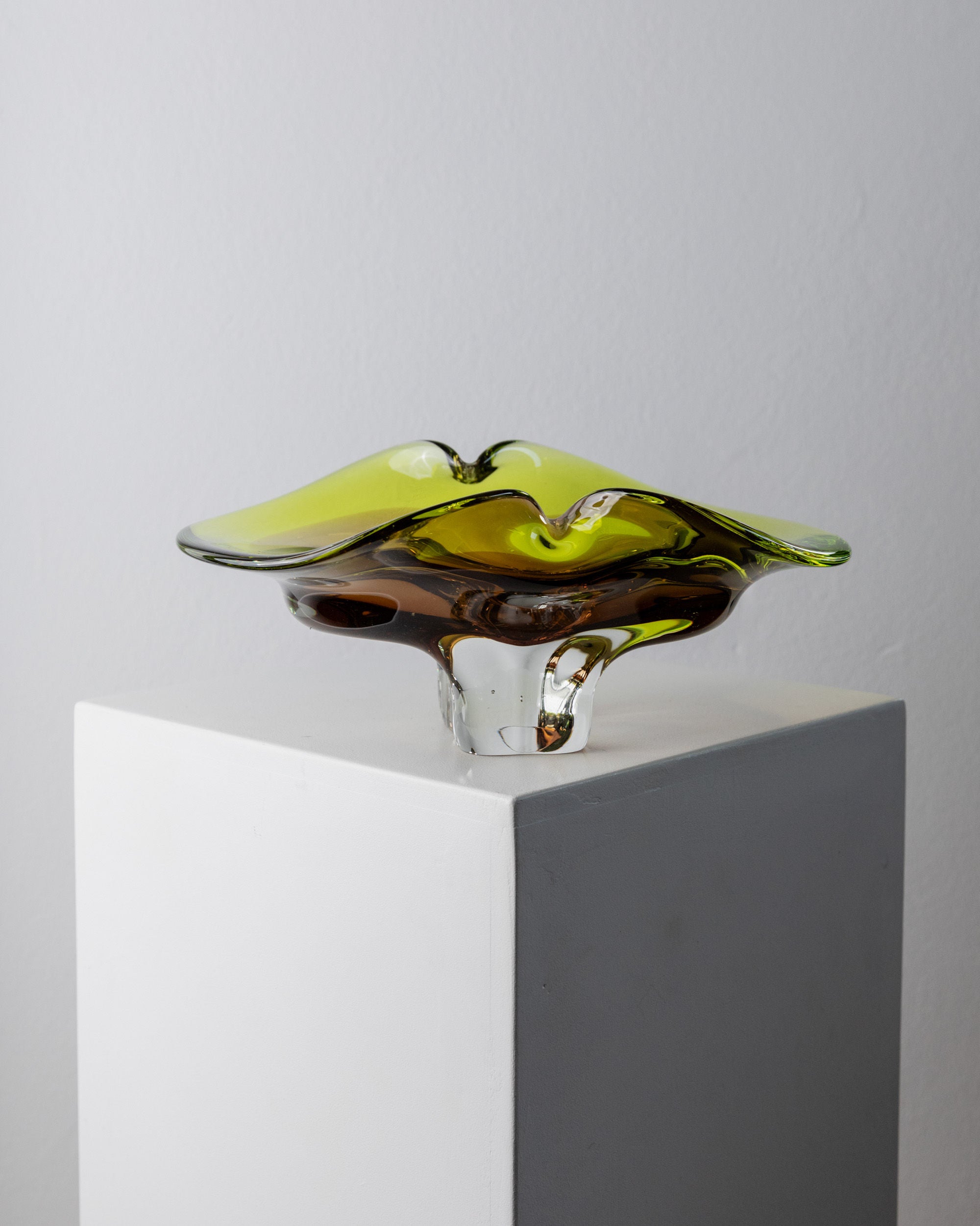 A Murano Glass Bowl from the 1970s by Bottega Jacobs, featuring yellow and clear sculpted glass with wavy edges, sits on a white pedestal, highlighting exquisite Italian craftsmanship against a plain background.