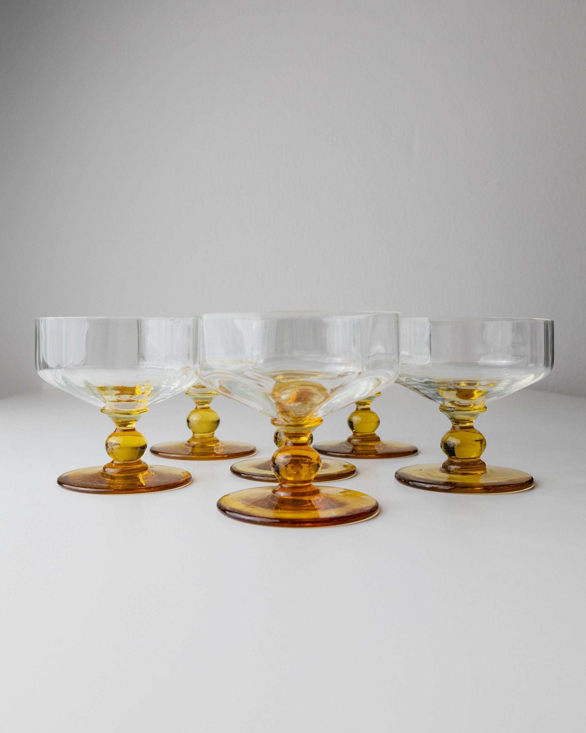 A set of six Bottega Jacobs Murano Glasses, Italy 70s, showcases their transparency and amber accents in a semi-circle on a white surface, evoking vintage charm through subtle color contrast against the plain background.