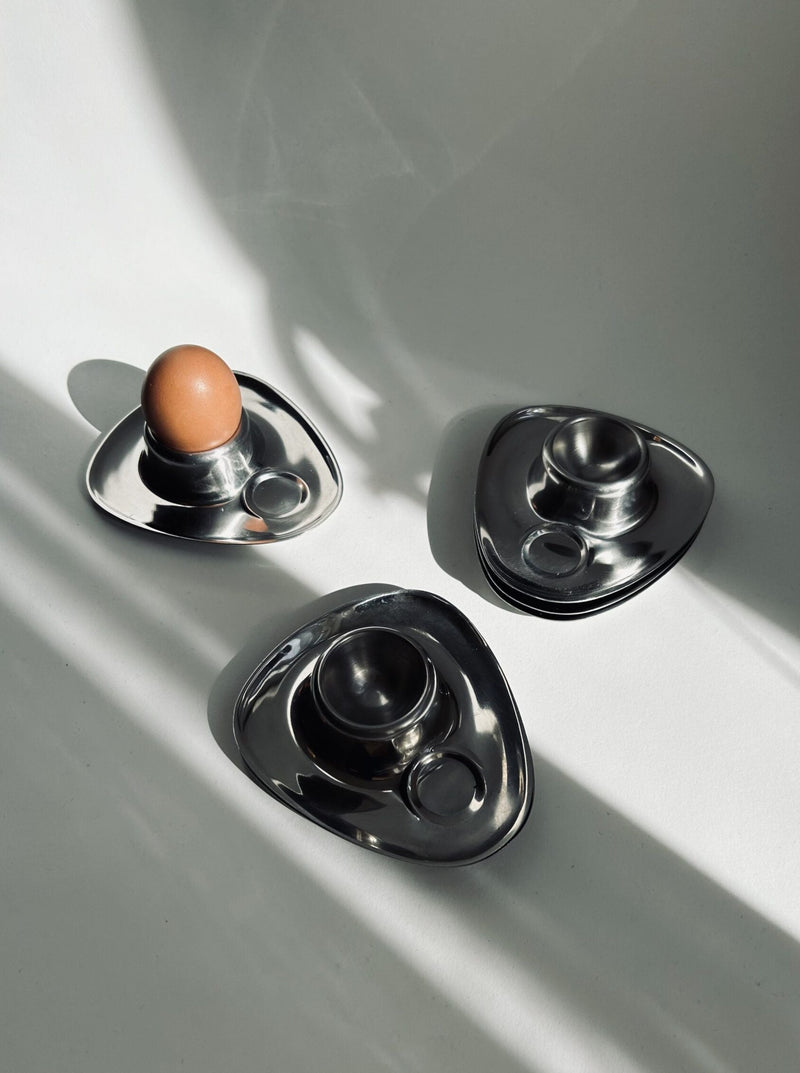 A trio of sleek, triangular egg holders from the Bauhaus Stainless Steel Egg Cups Set of 6 by Flock Studio are arranged on a white surface. One holder cradles a brown egg, while the others remain empty. Soft shadows stretch across the scene, adding depth and creating an elegant display reminiscent of vintage mid-century design.