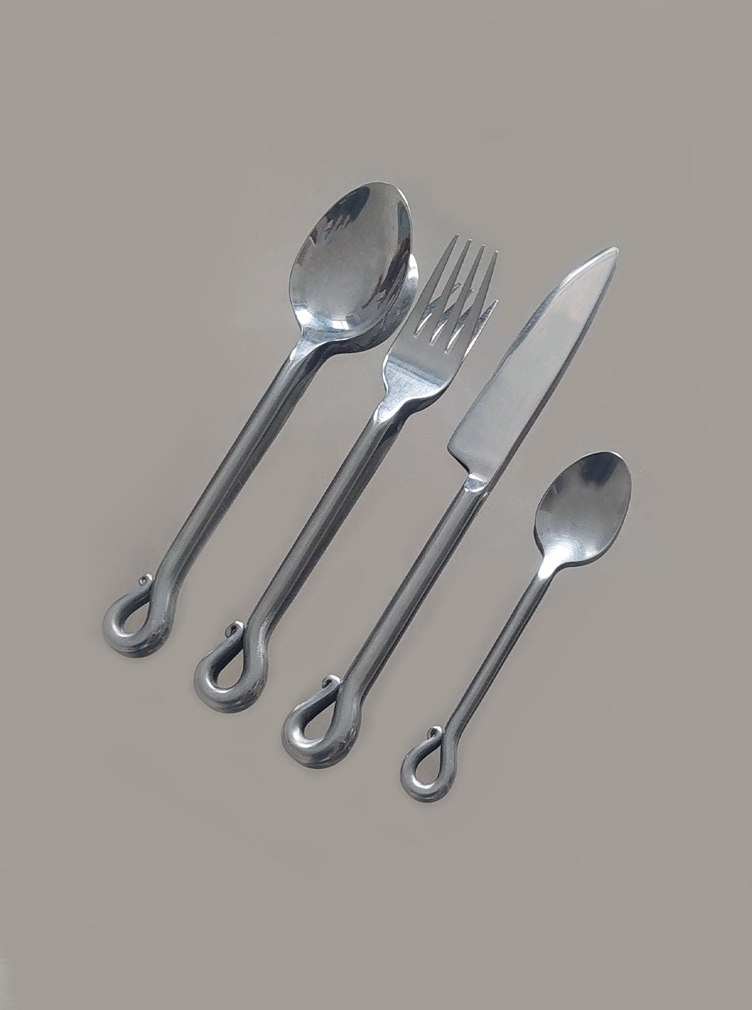 A set of four stunning stainless steel utensils from Les Objoies' Vintage Set of 4 Twist Cutlery artfully displayed on a light grey background. This elegant set, evocative of timeless cutlery design, includes a large spoon, a fork, a knife, and a small spoon. Each utensil is distinguished by its unique loop at the end of the handle.