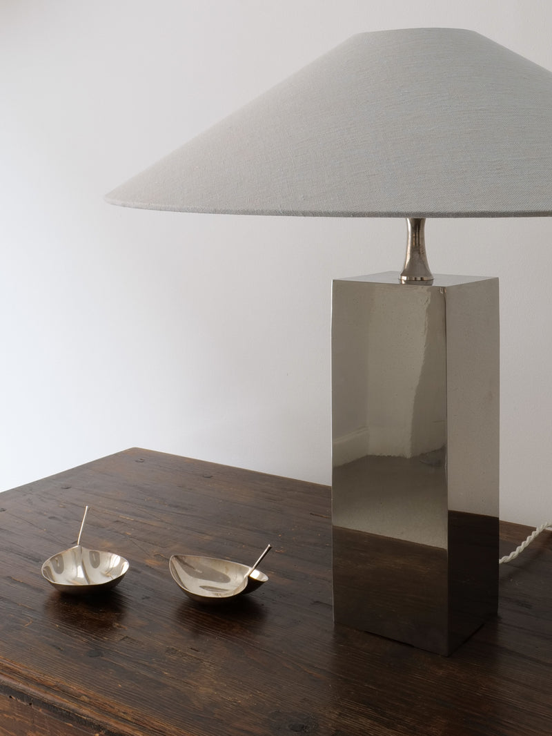 A modern, metallic table lamp with a rectangular base and a large, gray conical lampshade sits on a wooden surface. Two sleek Collection apart Tapio Wirkkala Silver Bowls with tiny handles are placed next to the lamp. The background features a plain white wall.
